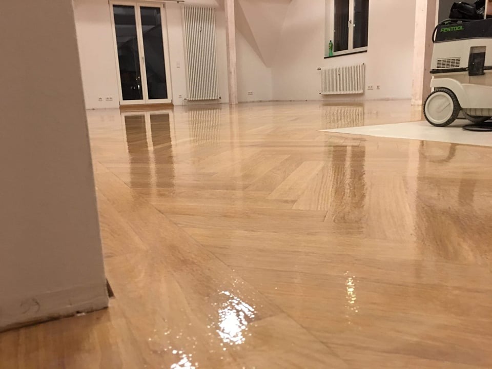 Błyszczący, świeżo polakierowany parkiet ułożony w jodełkę, odbijający światło okien. W tle widoczny odkurzacz przemysłowy i grzejniki.