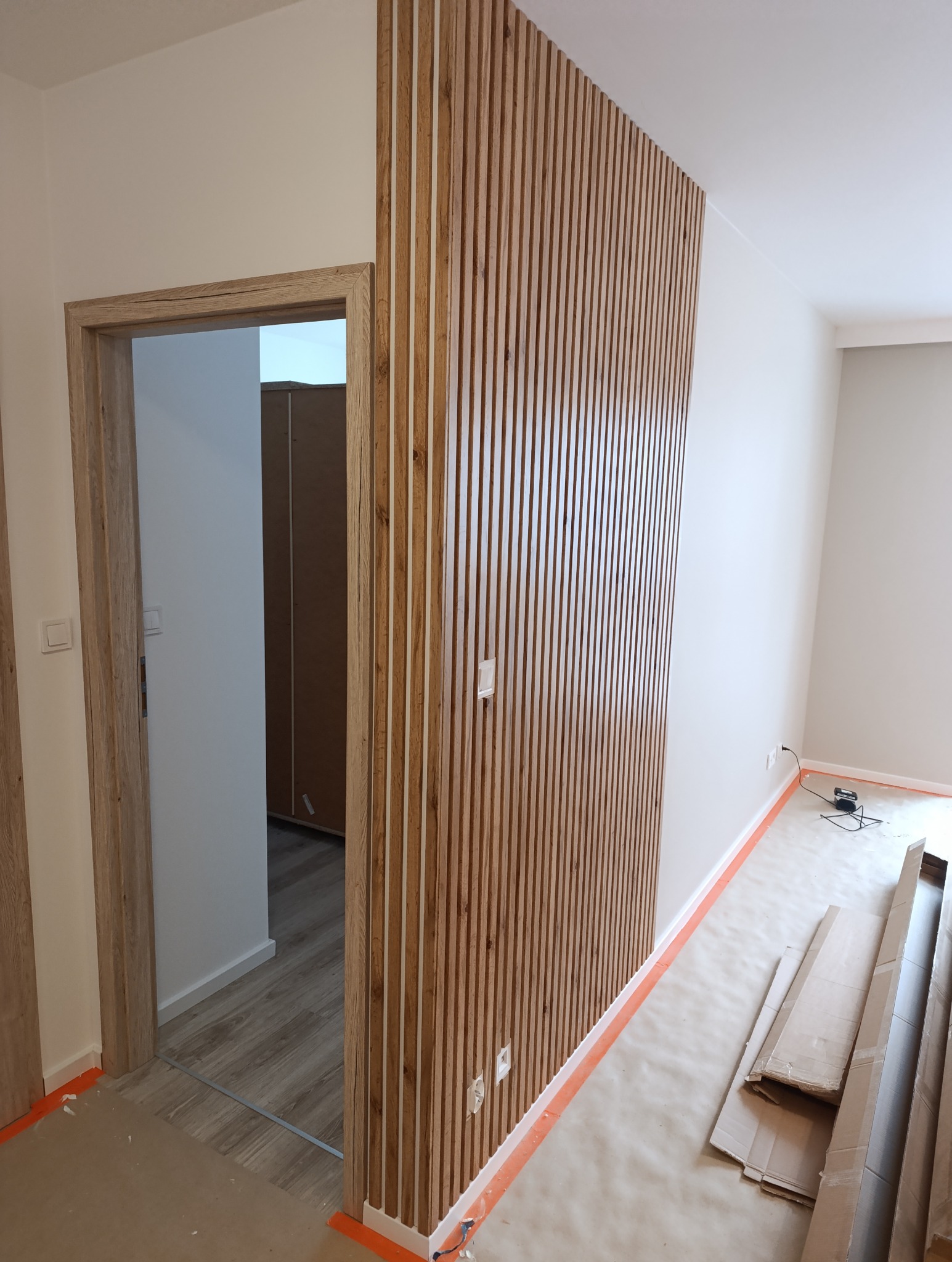 Drewniane lamele dekoracyjne na ścianie w jasnym wnętrzu, widoczne gniazdka elektryczne i fragment drzwi z ościeżnicą w podobnym odcieniu drewna. Podłoga zabezpieczona folią.