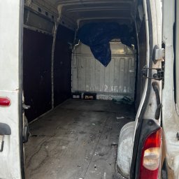 D.B Dariusz Błaszczyk - Wnętrze białego, pustego furgonetki z otwartymi drzwiami, widoczne ślady użytkowania na podłodze i ścianach, z akumulatorami i plandeką w tle.