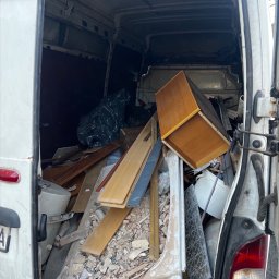 D.B Dariusz Błaszczyk - Wnętrze białego busa wypełnione gruzem, meblami i odpadami po remoncie. Widoczne deski, fragmenty płytek, szafka i czarny worek na śmieci. Otwarte drzwi pojazdu.