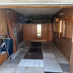 D.B Dariusz Błaszczyk - Garaż z drewnianą boazerią i szafami w zabudowie, częściowo opróżniony. Widoczna walizka, drabina i akcesoria. Podłoga wyłożona płytkami w różnych odcieniach szarości.