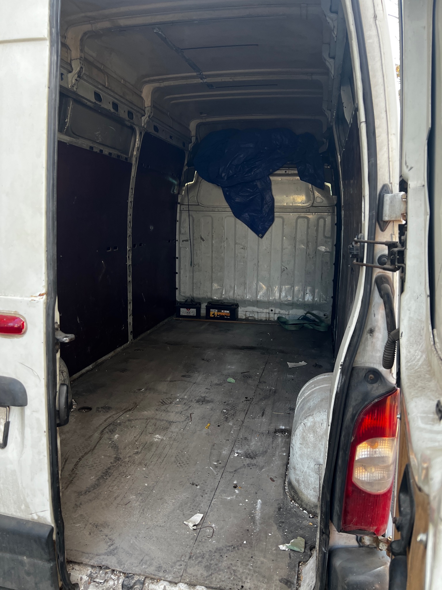 Wnętrze białego, pustego furgonetki z otwartymi drzwiami, widoczne ślady użytkowania na podłodze i ścianach, z akumulatorami i plandeką w tle.