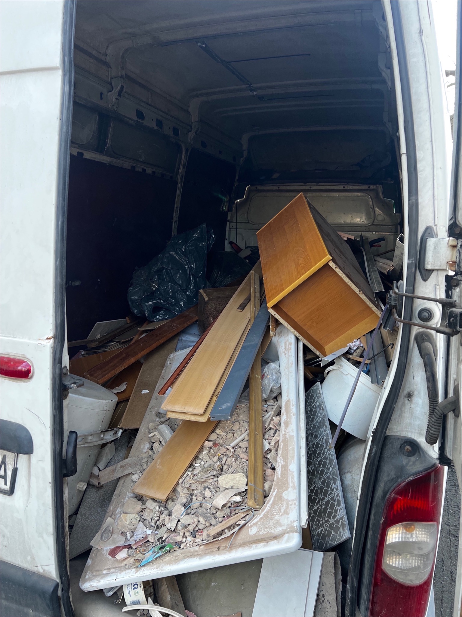 Wnętrze białego busa wypełnione gruzem, meblami i odpadami po remoncie. Widoczne deski, fragmenty płytek, szafka i czarny worek na śmieci. Otwarte drzwi pojazdu.