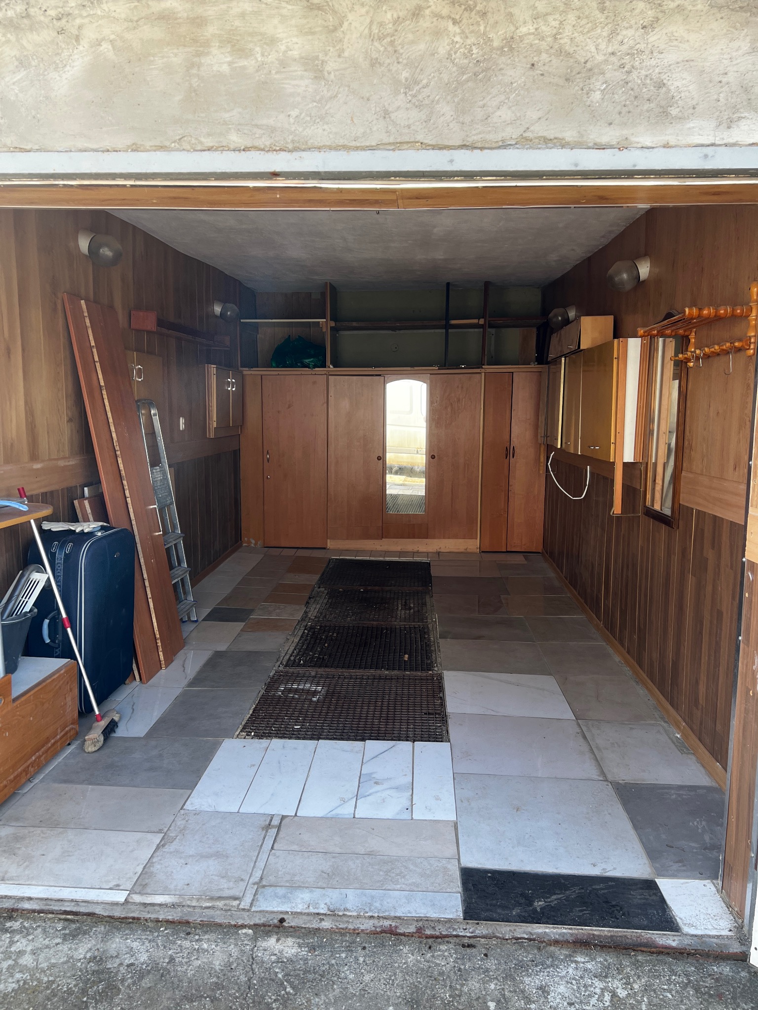 Garaż z drewnianą boazerią i szafami w zabudowie, częściowo opróżniony. Widoczna walizka, drabina i akcesoria. Podłoga wyłożona płytkami w różnych odcieniach szarości.