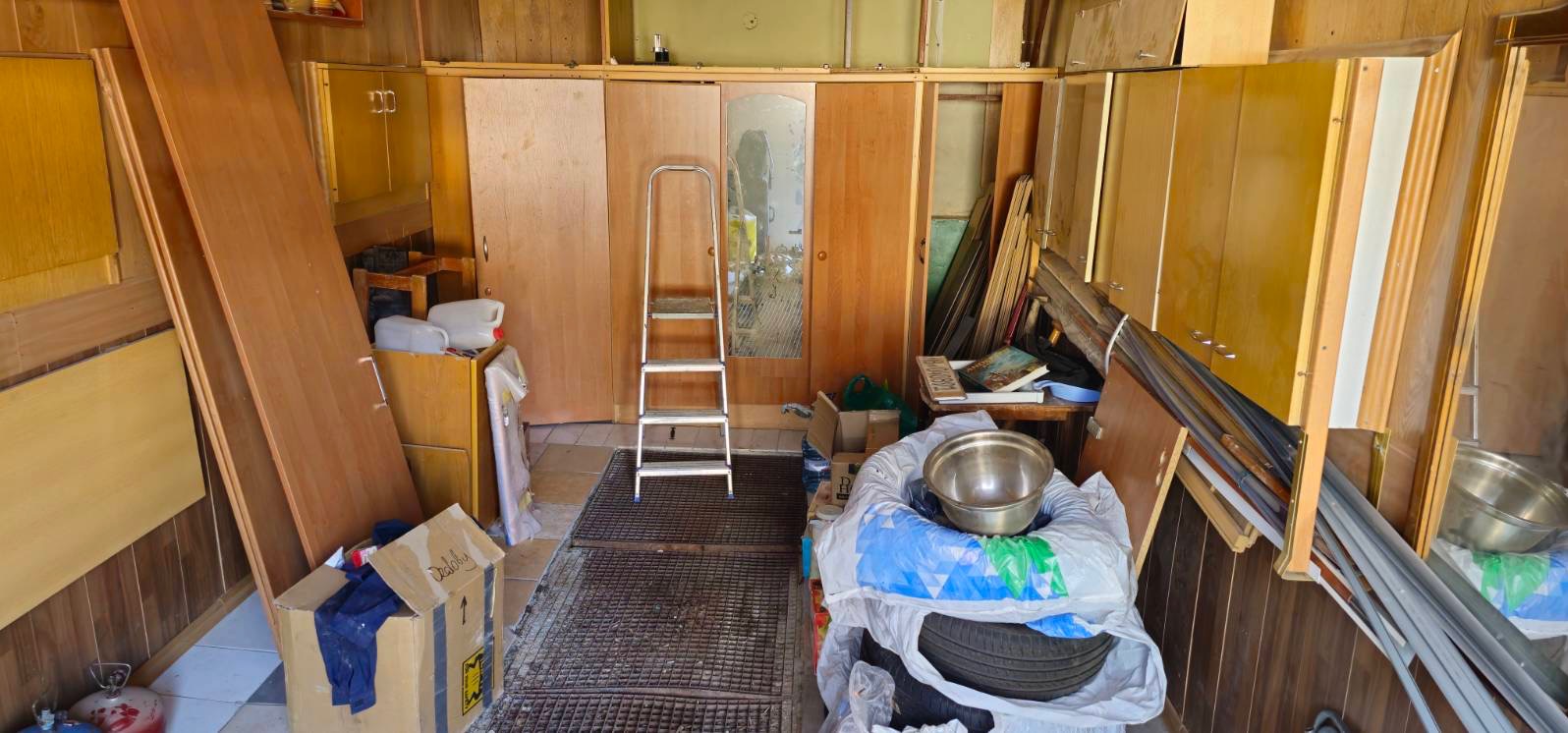 Zagracone pomieszczenie z drewnianą zabudową, kartonami, metalową drabinką i różnymi przedmiotami. Widoczne elementy wyposażenia przedmiotu.