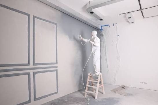 Pracownik w kombinezonie maluje ścianę z dekoracyjnymi ramkami za pomocą pistoletu natryskowego. Zabezpieczona folią część ściany. Praca na drabinie.