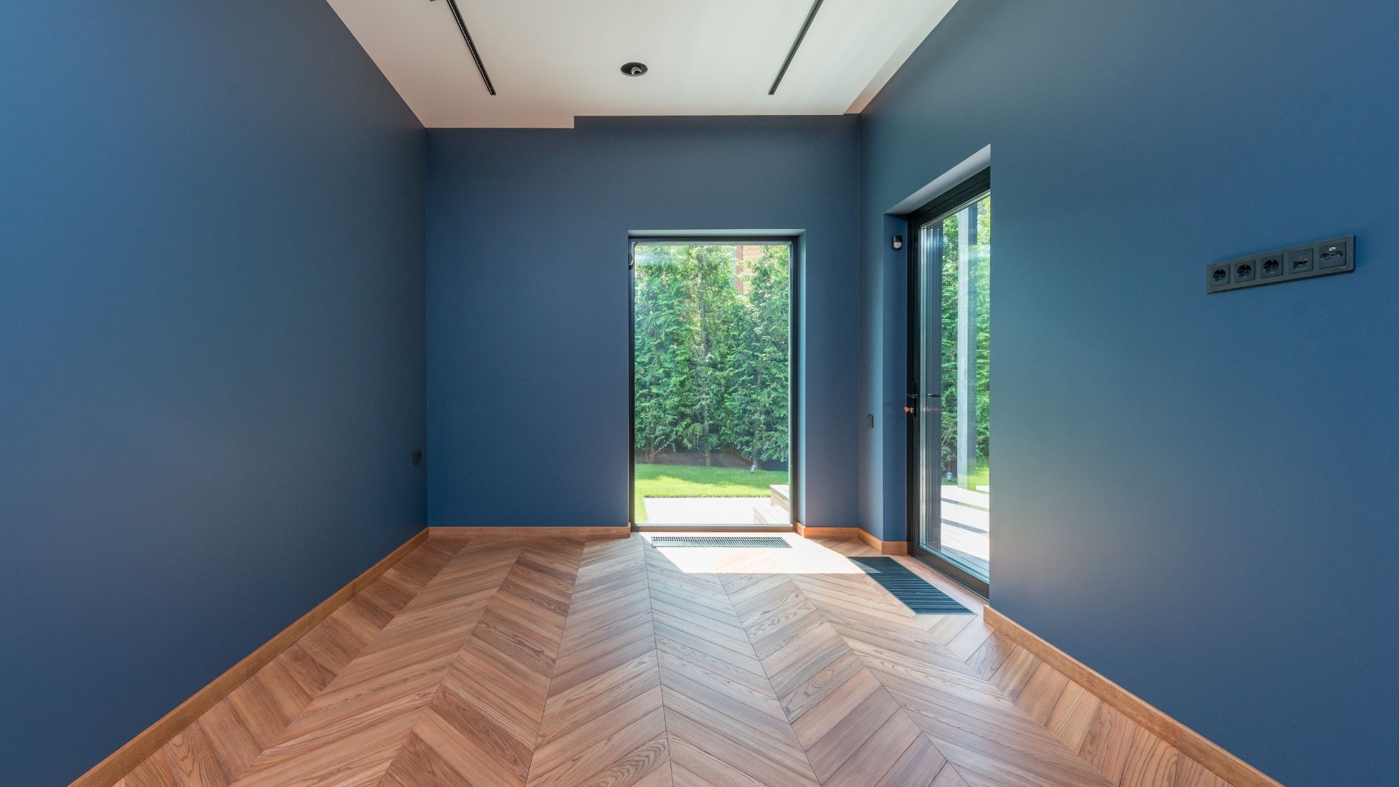 Błękitne ściany i parkiet w jodełkę w pustym pokoju z oknami na ogród. Gładkie ściany, nowoczesne gniazdka elektryczne. Minimalistyczne oświetlenie sufitowe.