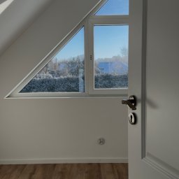 Krystian Sieradzki - Jasne poddasze z drewnianą podłogą, trójkątnym oknem z widokiem na zimowy krajobraz i otwartymi białymi drzwiami. W tle zwisająca żarówka.