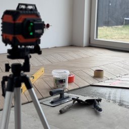 Krystian Sieradzki - Układanie parkietu w jodełkę, poziomica laserowa na statywie, narzędzia glazurnicze, wiaderko z dystansami. Częściowo ułożona podłoga w jasnym wnętrzu.
