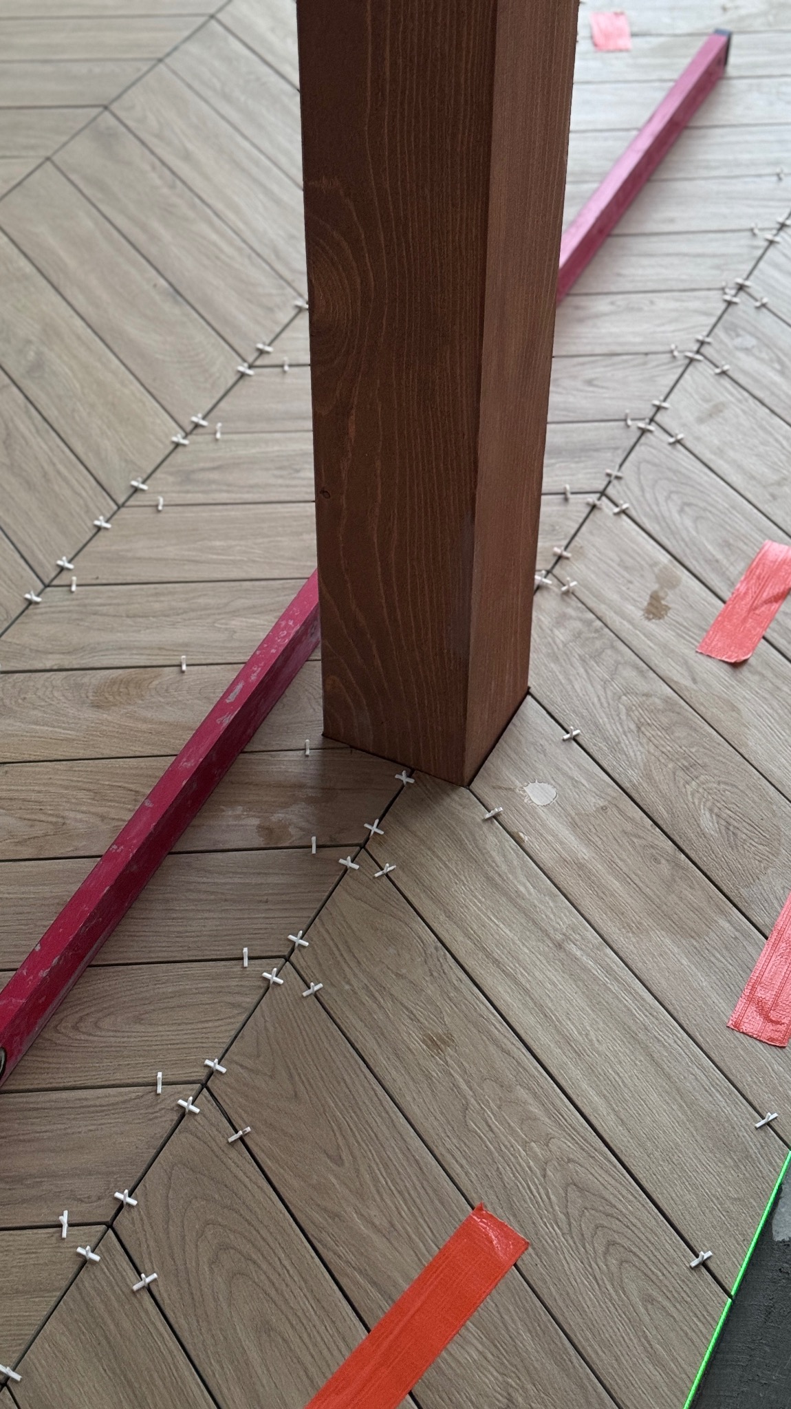 Ułożona pod kątem podłoga z płytek imitujących drewno, z krzyżykami dystansowymi i poziomicą, obok drewnianego słupka. Widoczne fragmenty taśmy malarskiej.