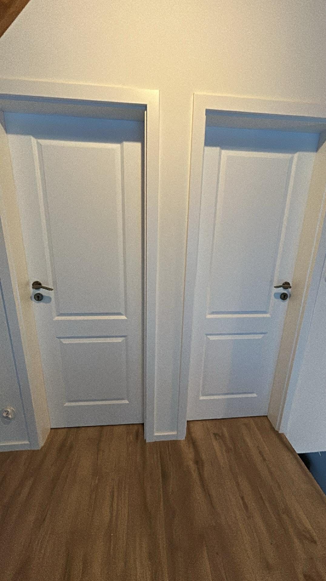 Dwa białe, identyczne drzwi wewnętrzne z klamkami, zamontowane obok siebie na tle drewnianej podłogi. Minimalistyczny design, symetryczny układ.