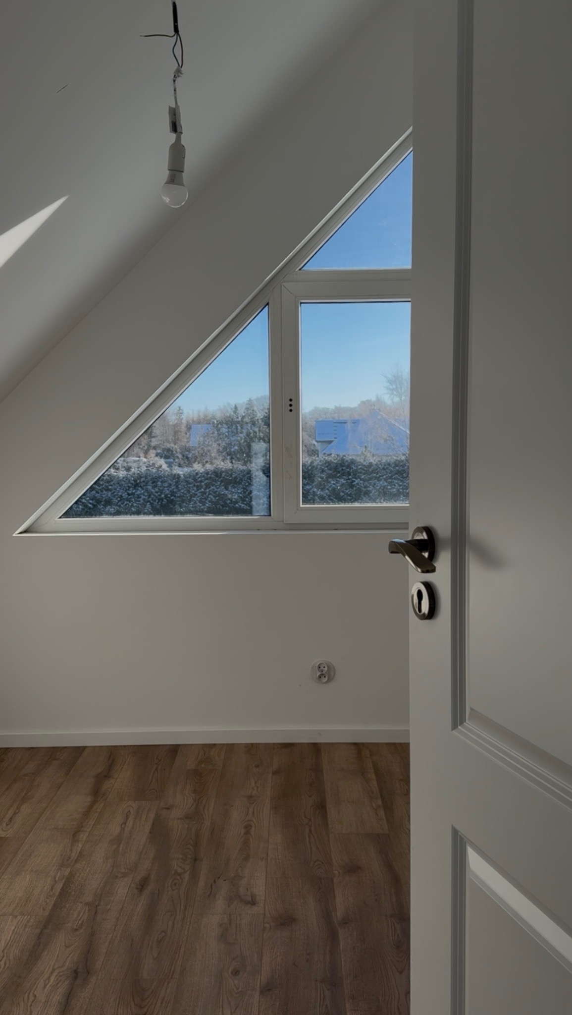 Jasne poddasze z drewnianą podłogą, trójkątnym oknem z widokiem na zimowy krajobraz i otwartymi białymi drzwiami. W tle zwisająca żarówka.