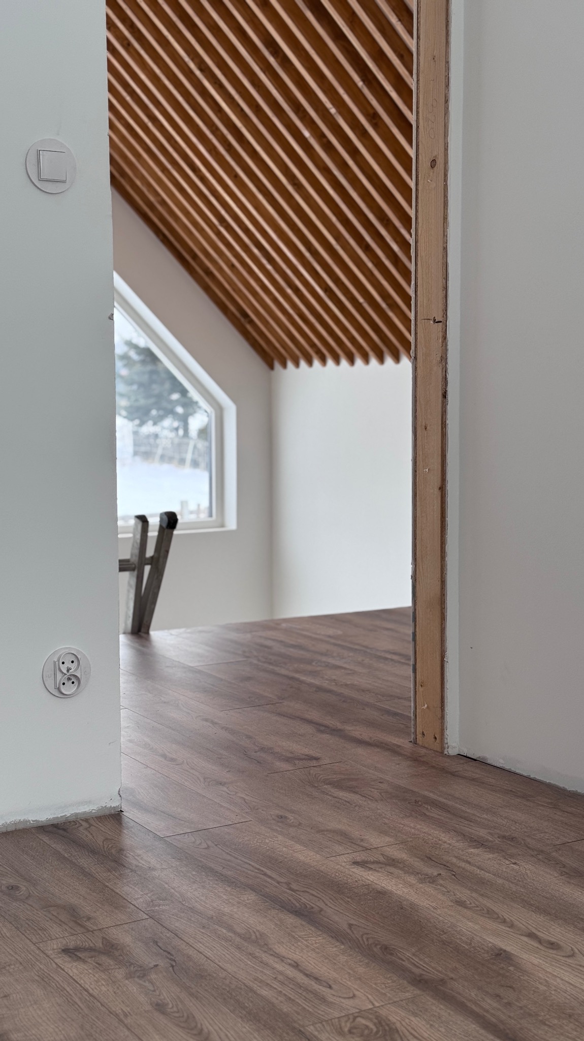 Ukończone układanie paneli podłogowych w Gdańsku, widok z perspektywy gniazdka elektrycznego, z drewnianym wykończeniem sufitu i oknem w tle.