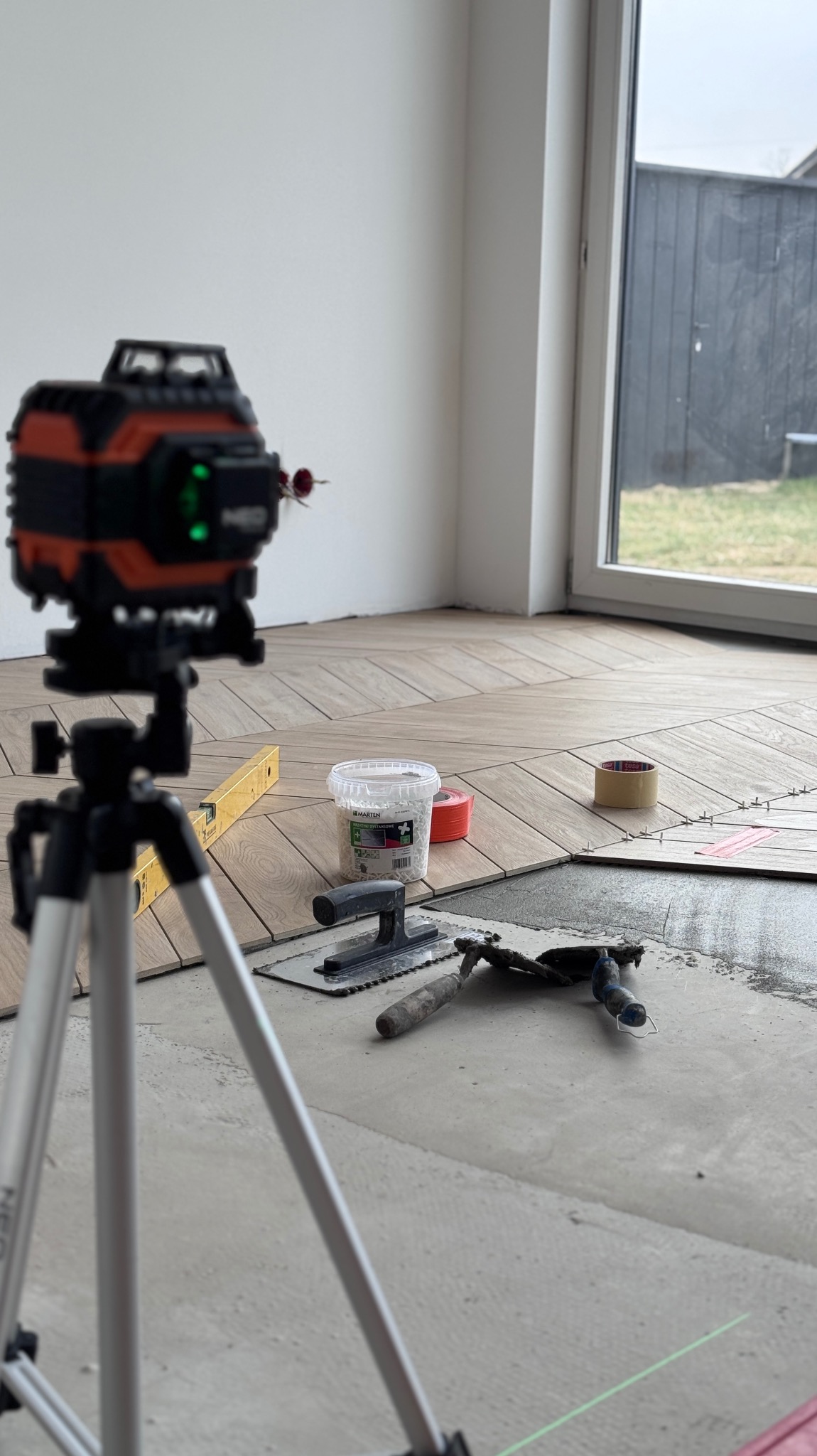 Układanie parkietu w jodełkę, poziomica laserowa na statywie, narzędzia glazurnicze, wiaderko z dystansami. Częściowo ułożona podłoga w jasnym wnętrzu.