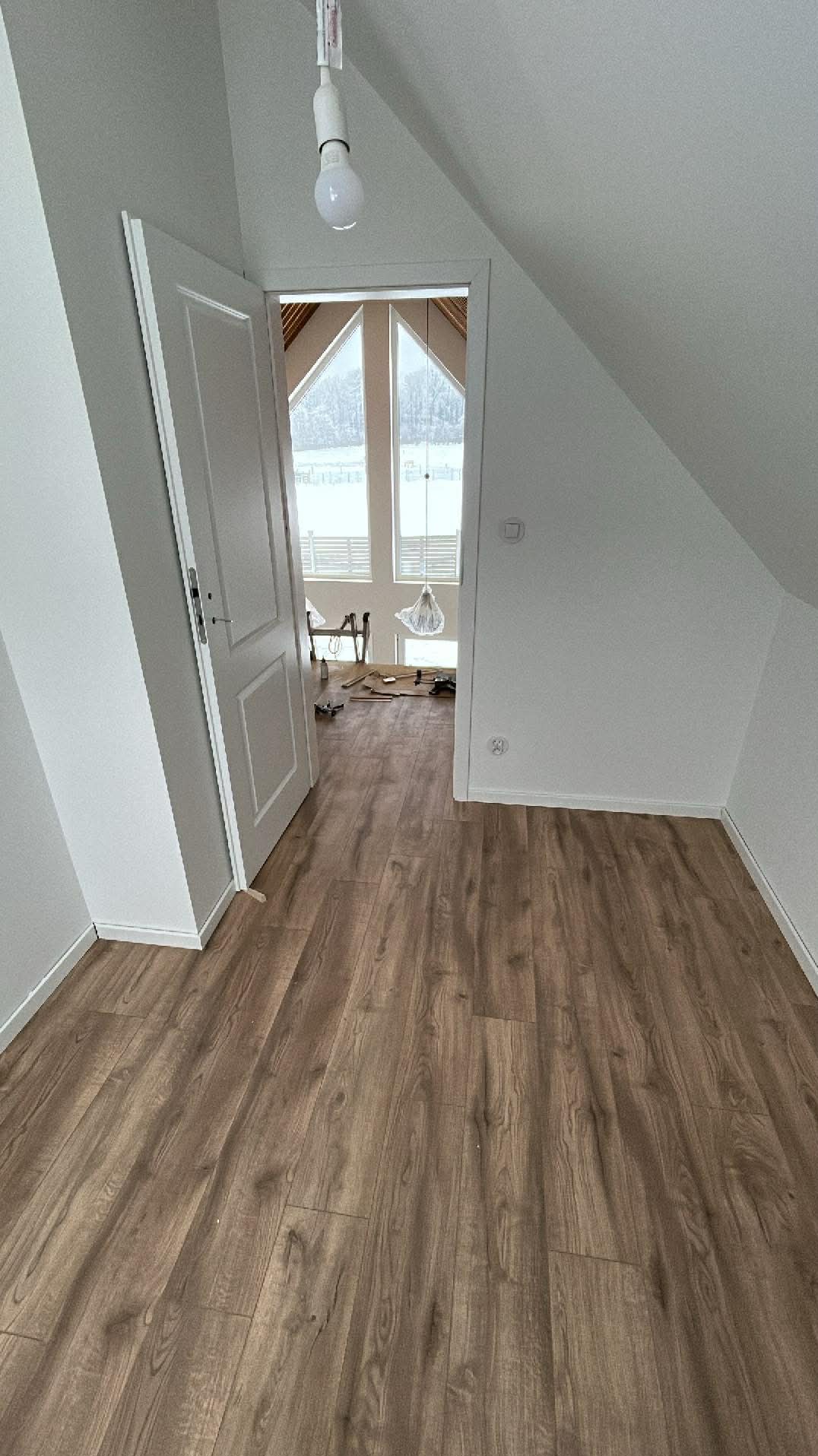 Jasne poddasze z drewnianą podłogą i otwartymi białymi drzwiami. W tle okna dachowe i narzędzia, sugerujące etap prac wykończeniowych.