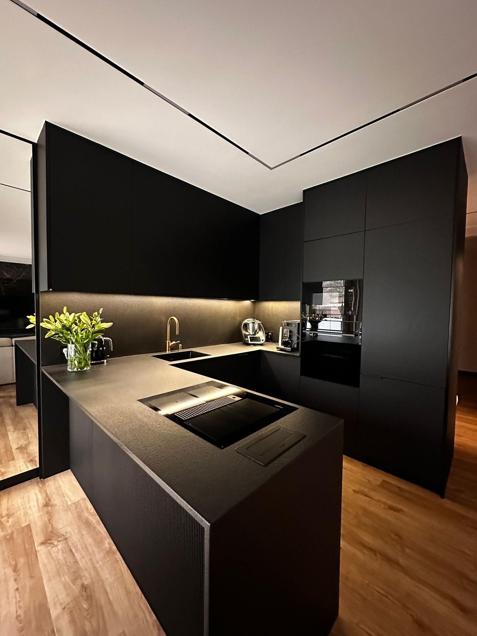 Nowoczesna, czarna kuchnia z wyspą i drewnianą podłogą. Minimalistyczny design z oświetleniem LED i złotą armaturą. Widoczne sprzęty AGD i dekoracyjny bukiet.