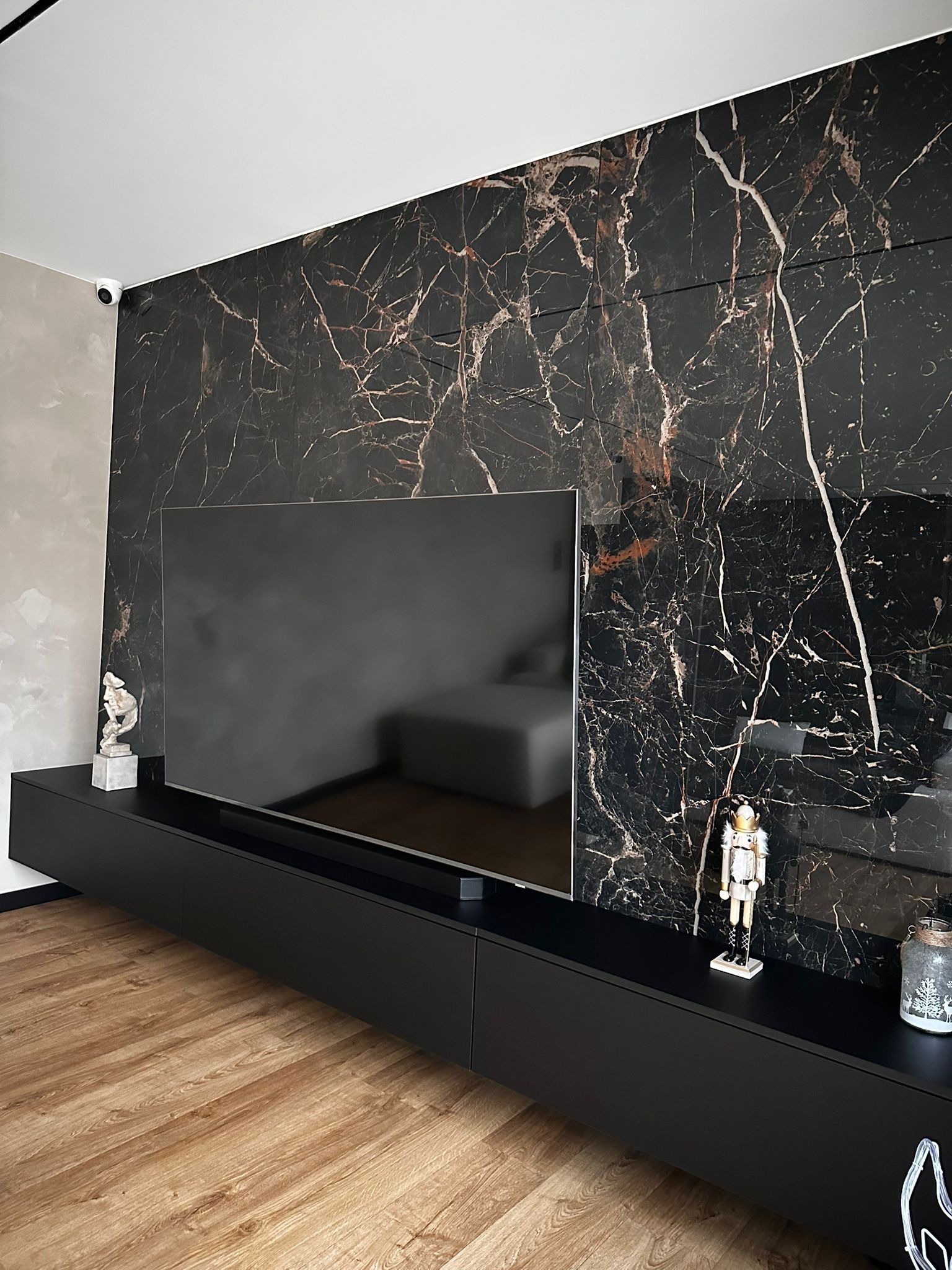 Salon z czarną ścianą z efektem marmuru, podświetlonym telewizorem i niską, czarną szafką RTV z dekoracjami. Parkiet na podłodze.