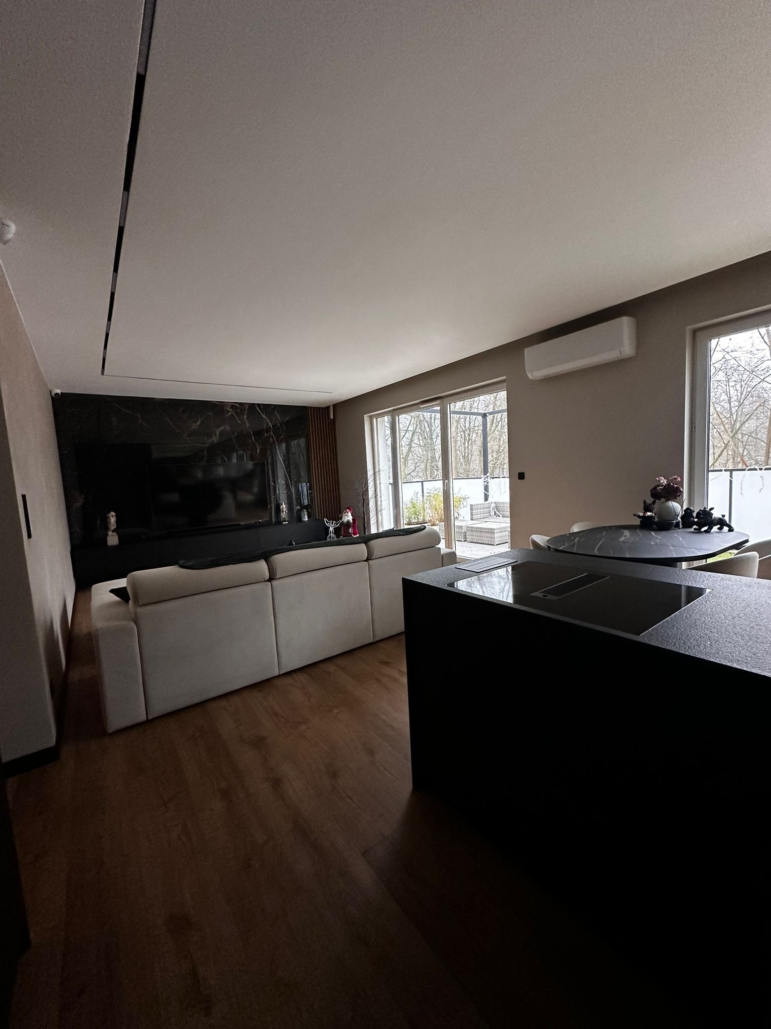 Nowoczesny salon z aneksem kuchennym: beżowa sofa, czarny panel TV z marmurowym wzorem, okrągły stół i wyspa kuchenna z płytą. Widok na taras przez przesuwne drzwi.