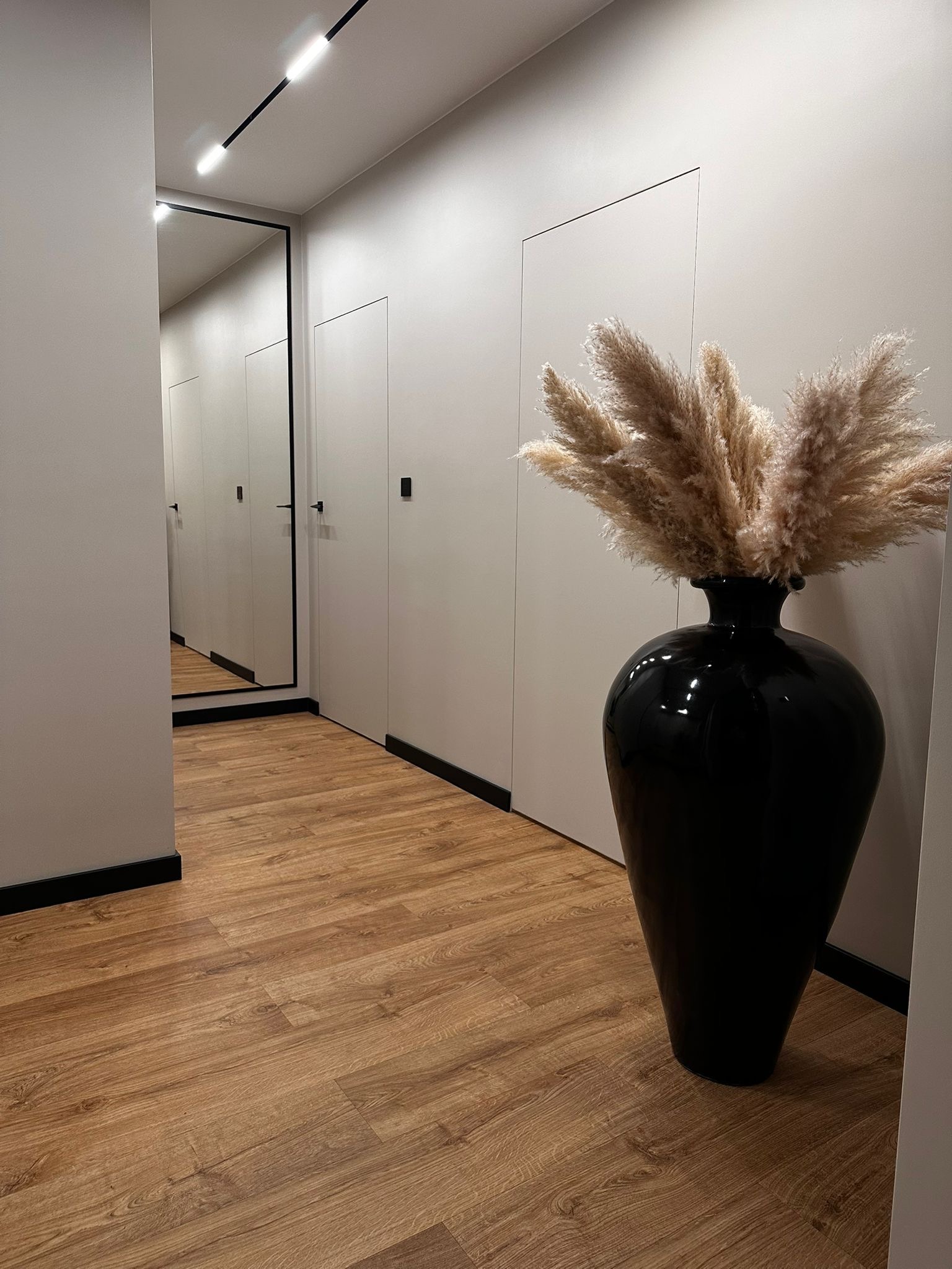 Korytarz z drewnianą podłogą, białymi ścianami i ukrytymi drzwiami. Duże czarne lustro i wazon z pampasową trawą dodają elegancji.