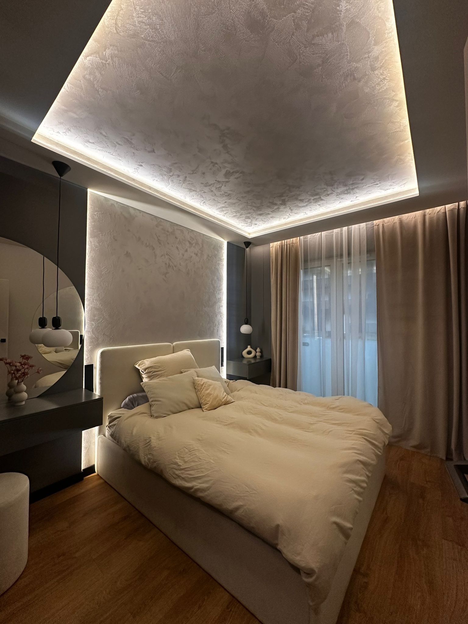 Nowoczesna sypialnia z podświetlanym sufitem i ścianą, okrągłym lustrem nad toaletką i beżową pościelą. Minimalistyczny design z akcentami światła.