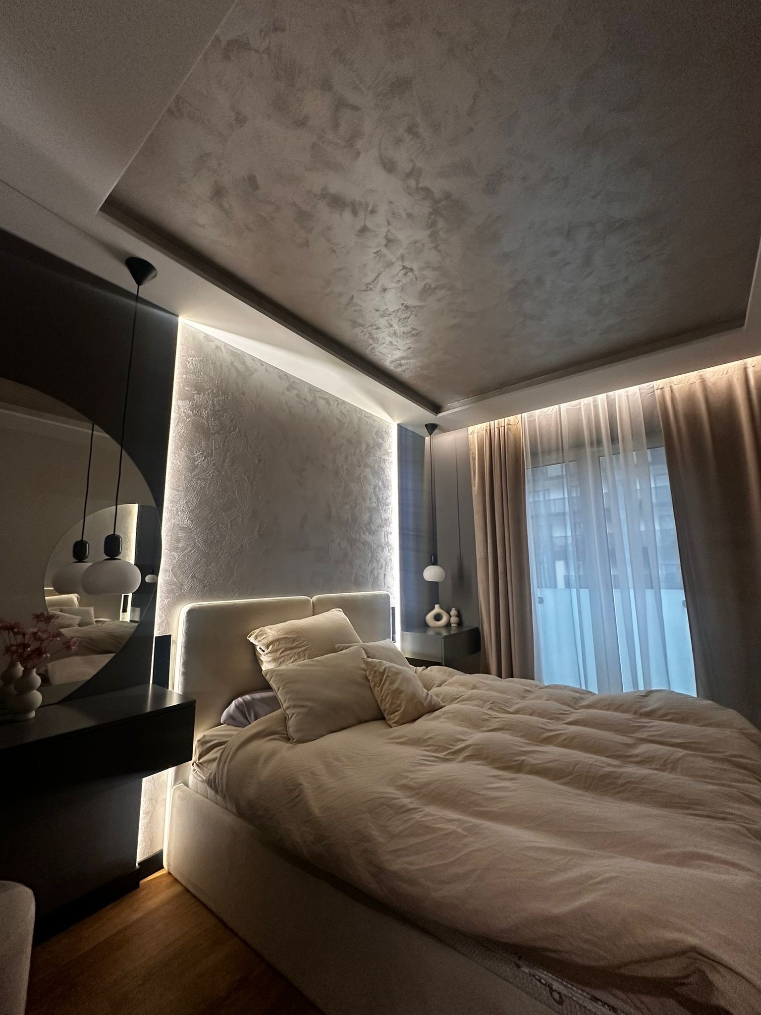 Nowoczesna sypialnia z łóżkiem w beżowej pościeli, okrągłym lustrem i oświetleniem LED. Dekoracyjna faktura na suficie i ścianie. Minimalistyczny design.