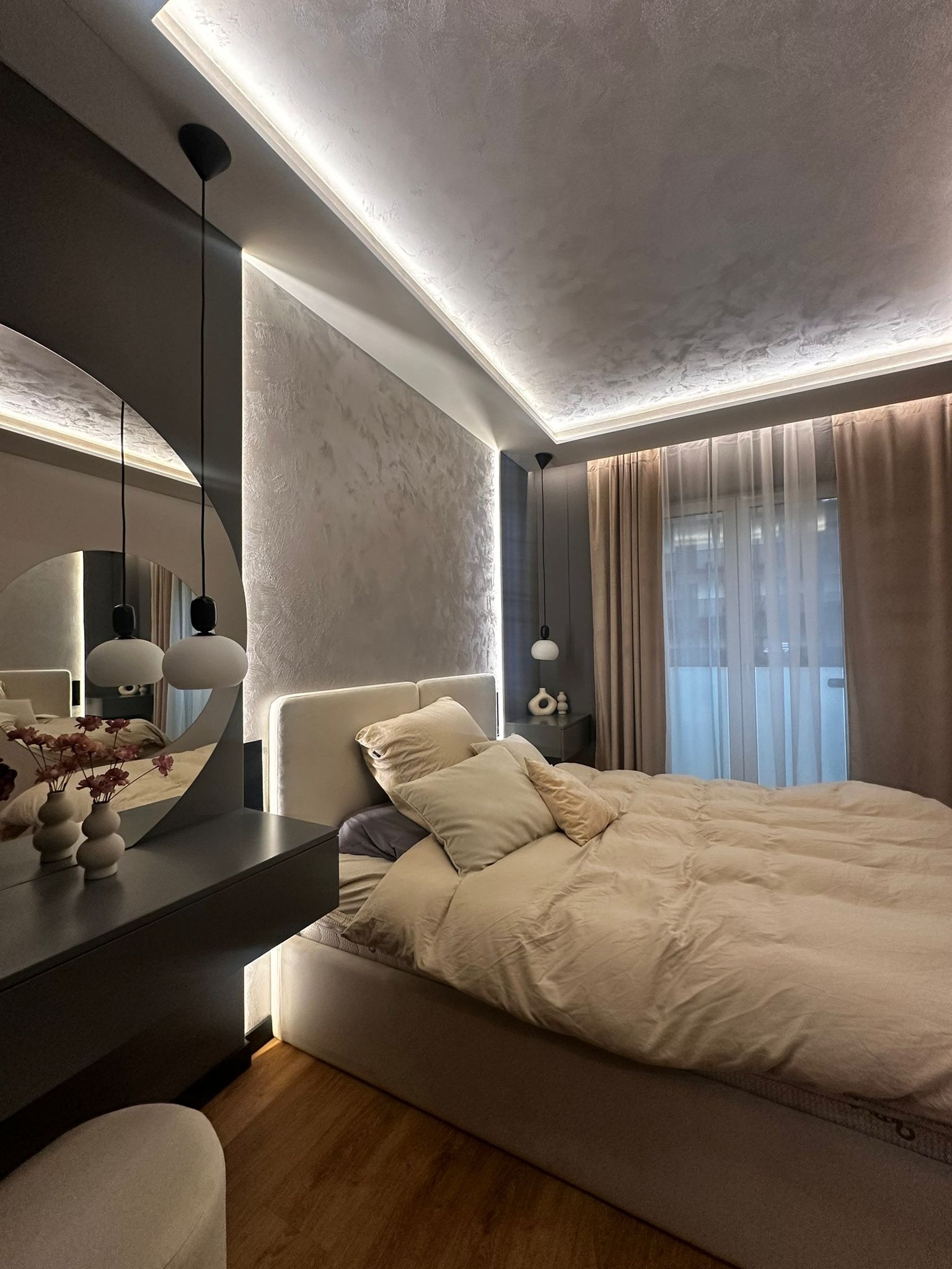 Nowoczesna sypialnia z łóżkiem w beżowej pościeli, okrągłym lustrem nad toaletką i subtelnym oświetleniem LED na suficie i pod meblami. Minimalistyczny design.