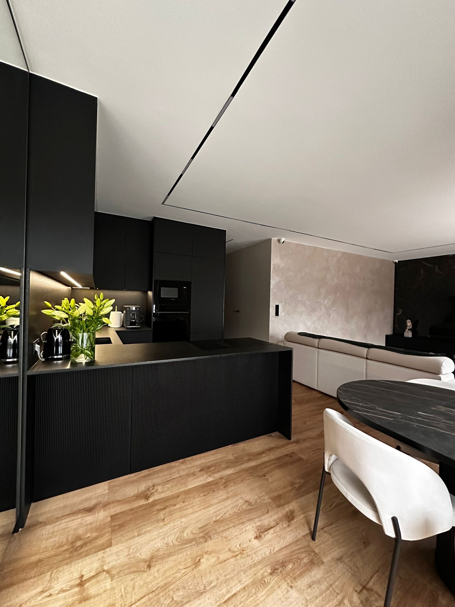 Nowoczesna kuchnia z czarnymi szafkami, wyspą z ryflowanym frontem i drewnianą podłogą. W tle salon z beżową sofą i dekoracyjną ścianą.