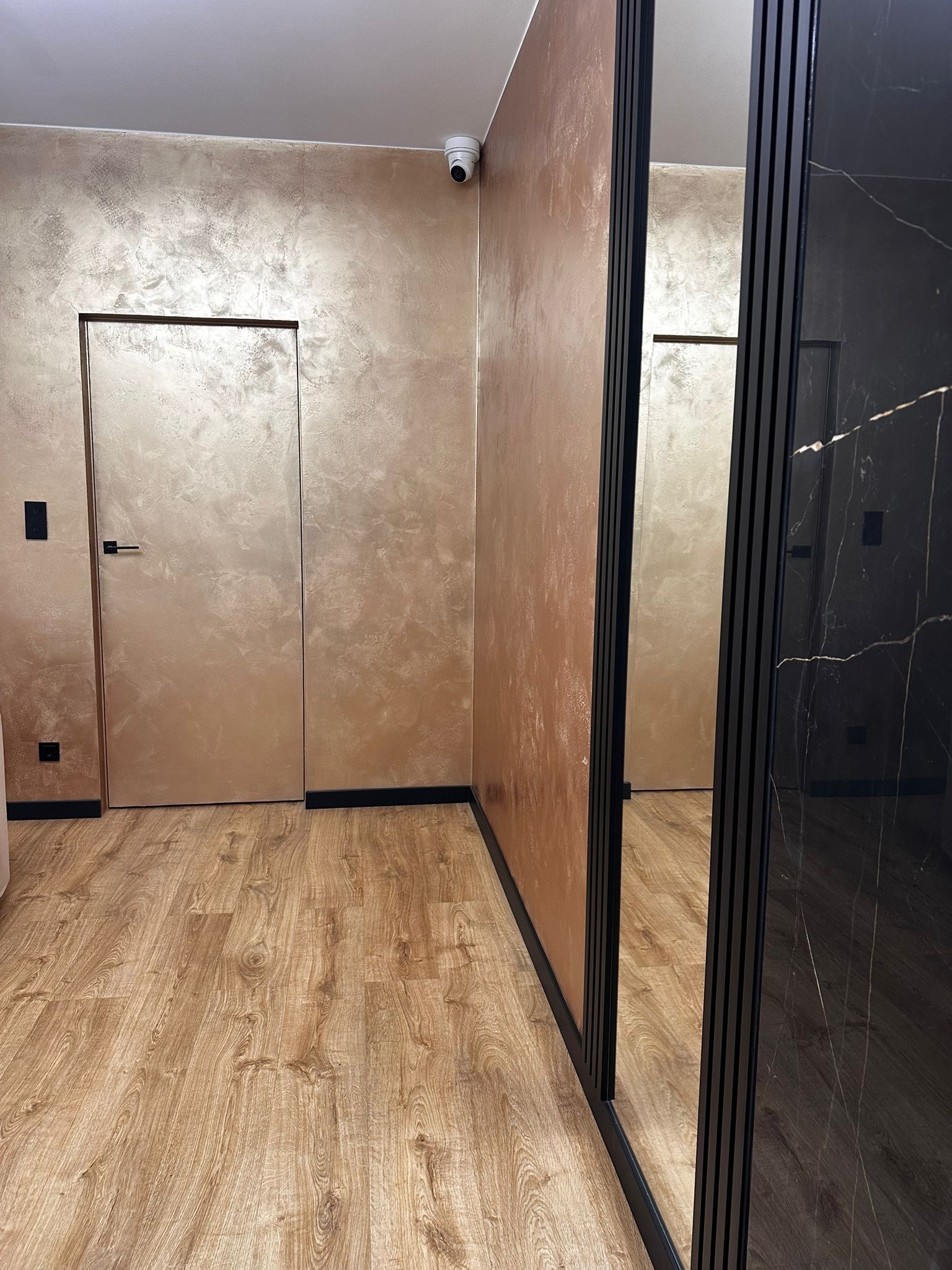 Wnętrze z drewnianą podłogą, ścianami w odcieniach złota i miedzi oraz szafą z czarnym marmurem. Widoczny fragment drzwi i kamera bezpieczeństwa.