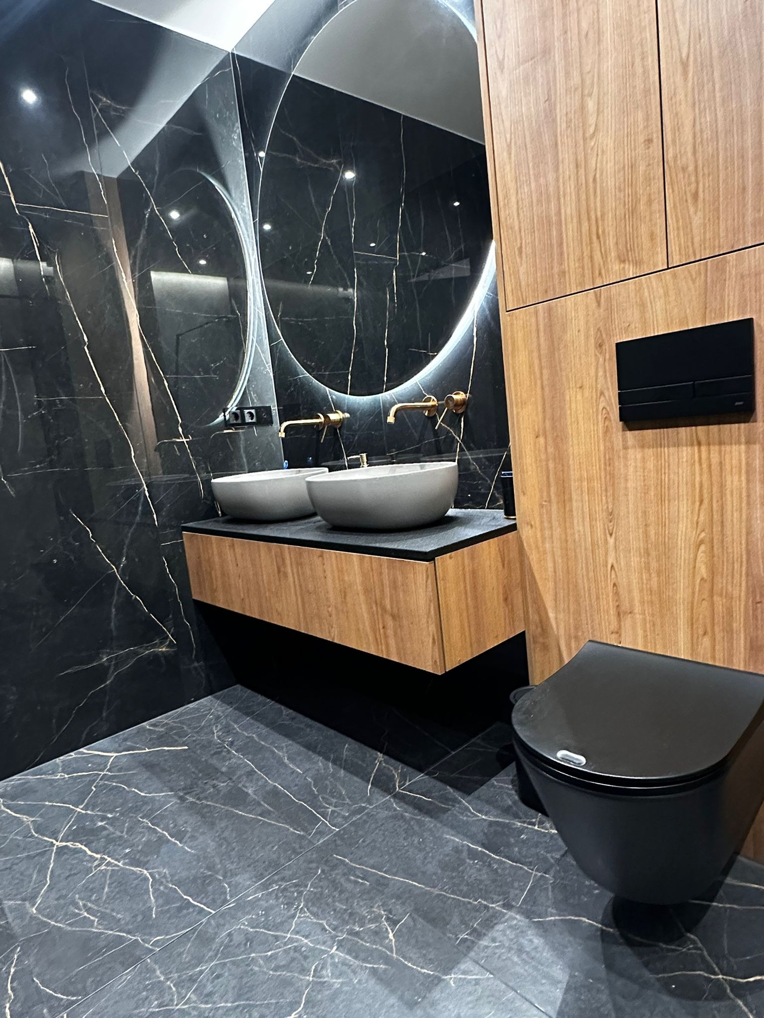 Nowoczesna łazienka z czarnymi marmurowymi płytkami, owalnym lustrem z podświetleniem LED, dwiema umywalkami nablatowymi i drewnianą szafką pod umywalką.