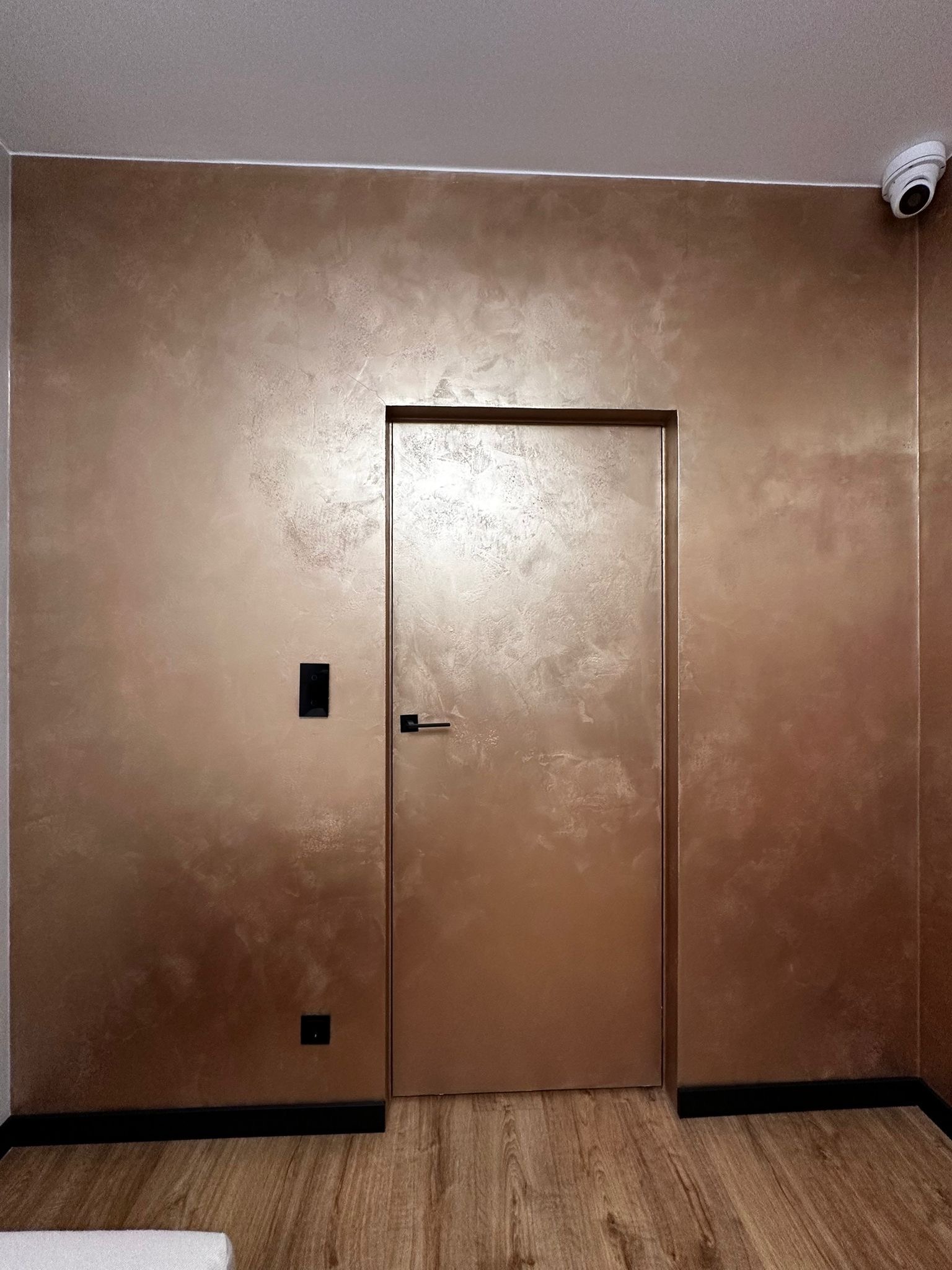 Ściana i ukryte drzwi w odcieniach brązu z efektem dekoracyjnym, czarne listwy przypodłogowe i drewniana podłoga. W rogu widoczna kamera.