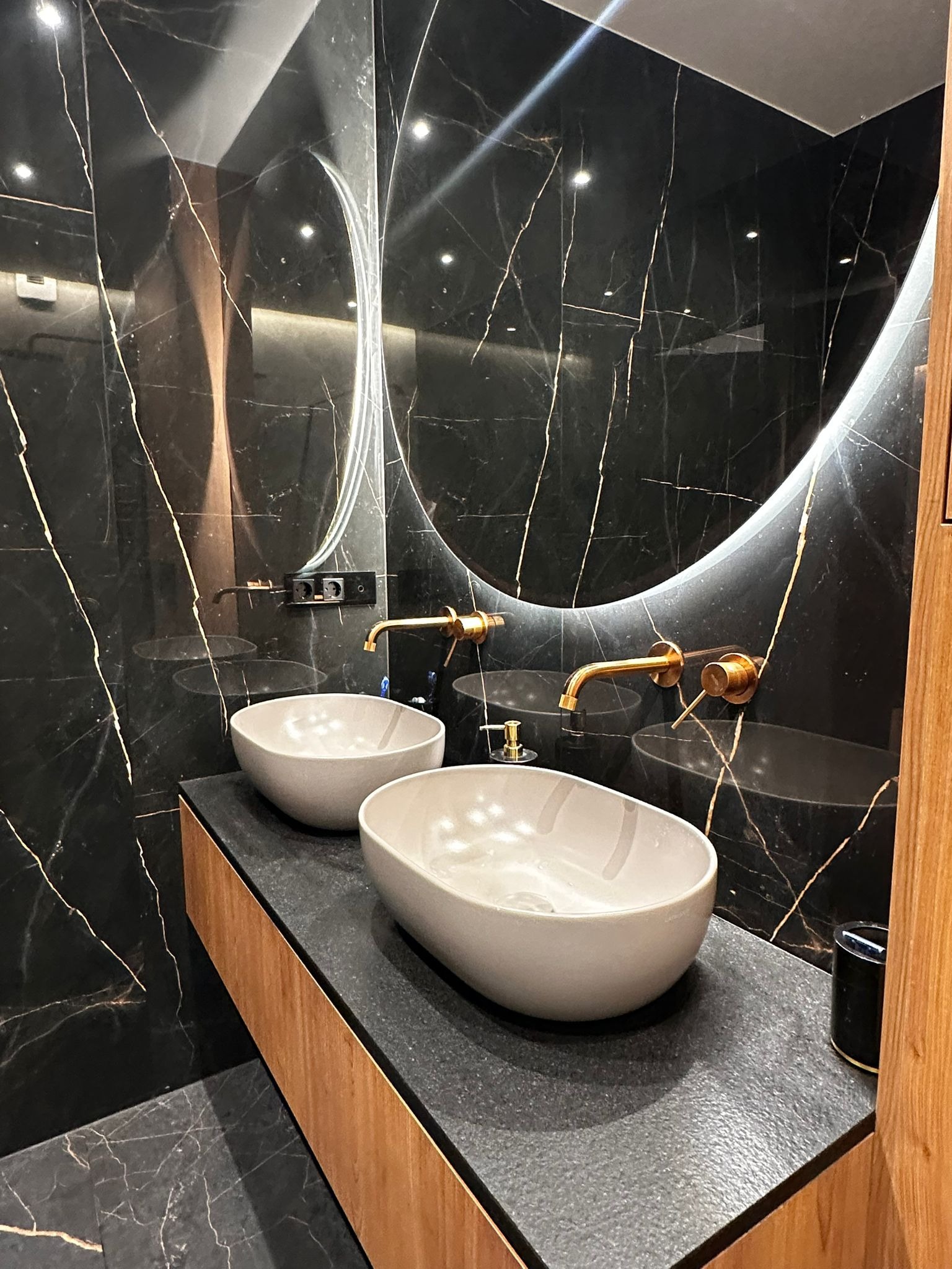 Elegancka łazienka z czarnym marmurem, złotą armaturą i owalnymi umywalkami. Okrągłe lustro z podświetleniem LED dodaje nowoczesnego charakteru.