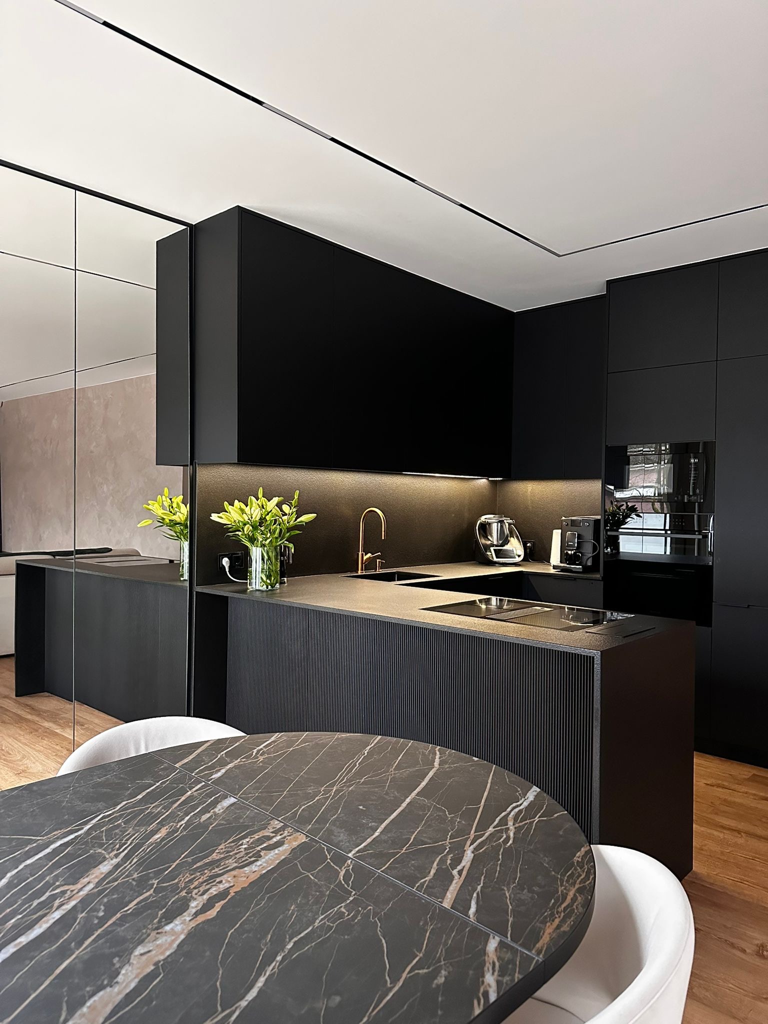 Nowoczesna, czarna kuchnia z wyspą i marmurowym stołem. Minimalistyczny design, oświetlenie LED pod szafkami, drewniana podłoga. Widok na lustrzaną ścianę.