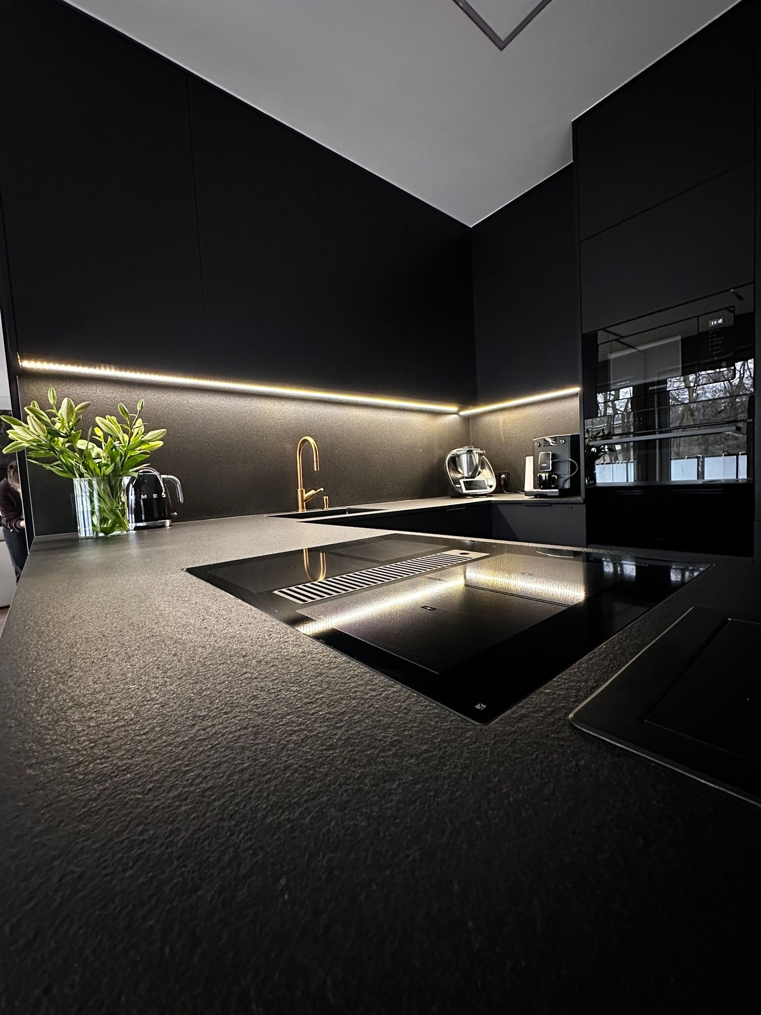 Nowoczesna, czarna kuchnia z kamiennym blatem, złotą armaturą i subtelnym oświetleniem LED. Minimalistyczny design z eleganckimi akcentami i wysokiej jakości sprzętem.