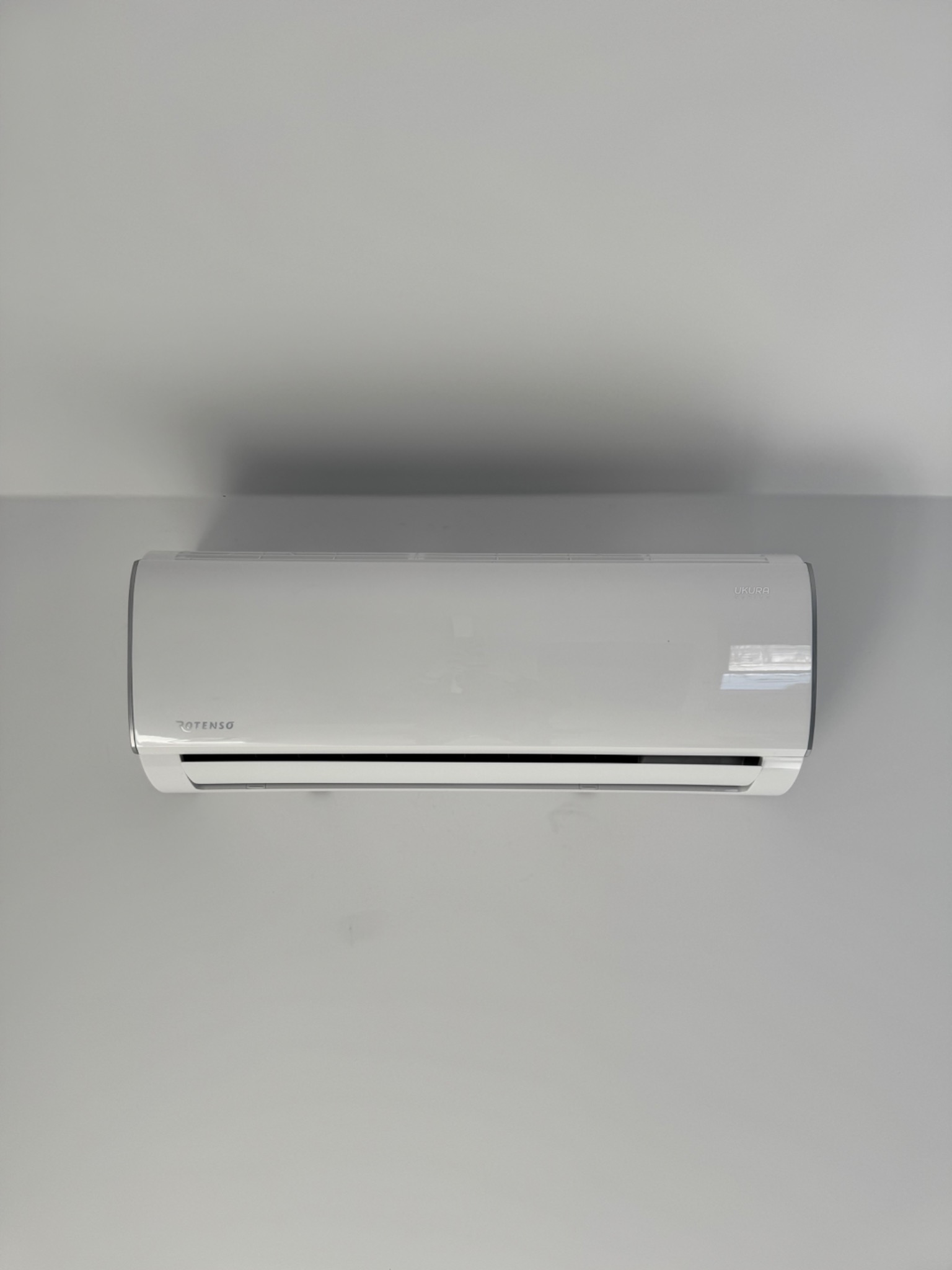 Biały klimatyzator Rotenso Ukura zamontowany na szarej ścianie, widok z przodu, minimalistyczny design, czyste linie, nowoczesne wnętrze.