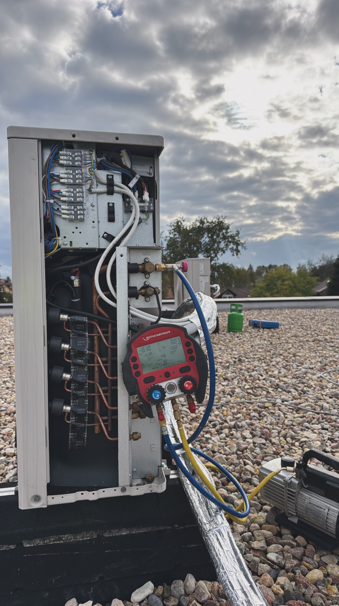 Otwarta obudowa klimatyzatora na dachu z podłączonym manometrem cyfrowym Rothenberger i pompą próżniową. Widoczne przewody i złącza. Pochmurne niebo w tle.
