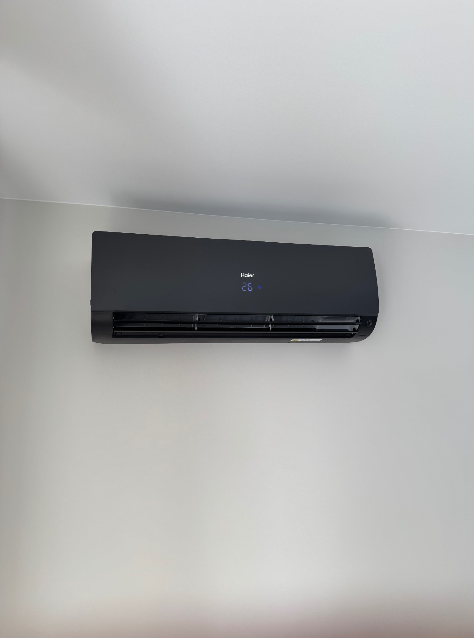 Czarny klimatyzator Haier zamontowany na jasnej ścianie. Wyświetlacz pokazuje temperaturę 26 stopni. Minimalistyczny design, nowoczesne wnętrze.