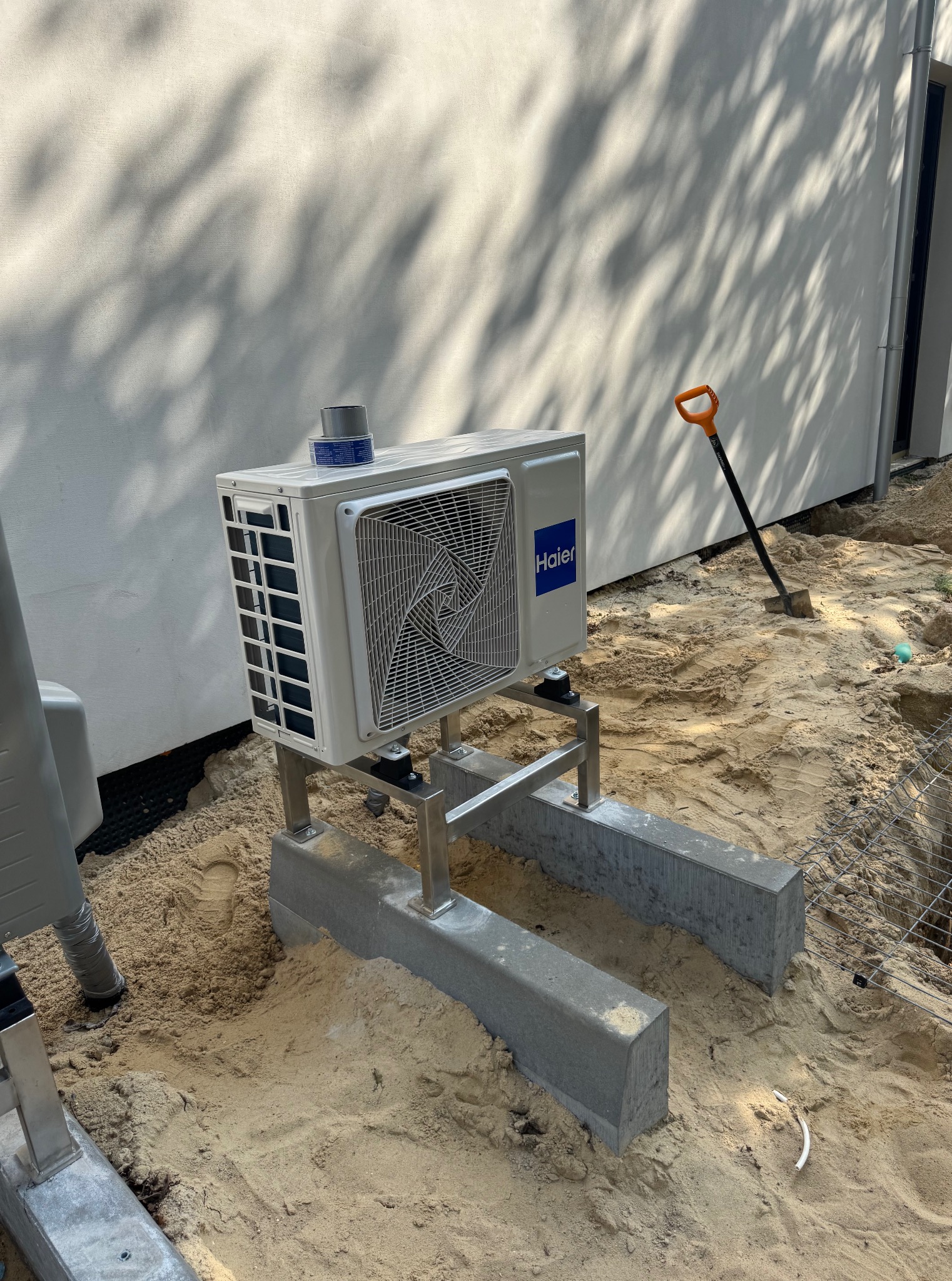 Biała jednostka zewnętrzna klimatyzacji Haier, zamontowana na betonowych podstawach i metalowej konstrukcji, na piaszczystym podłożu. W tle biała ściana budynku i łopata.