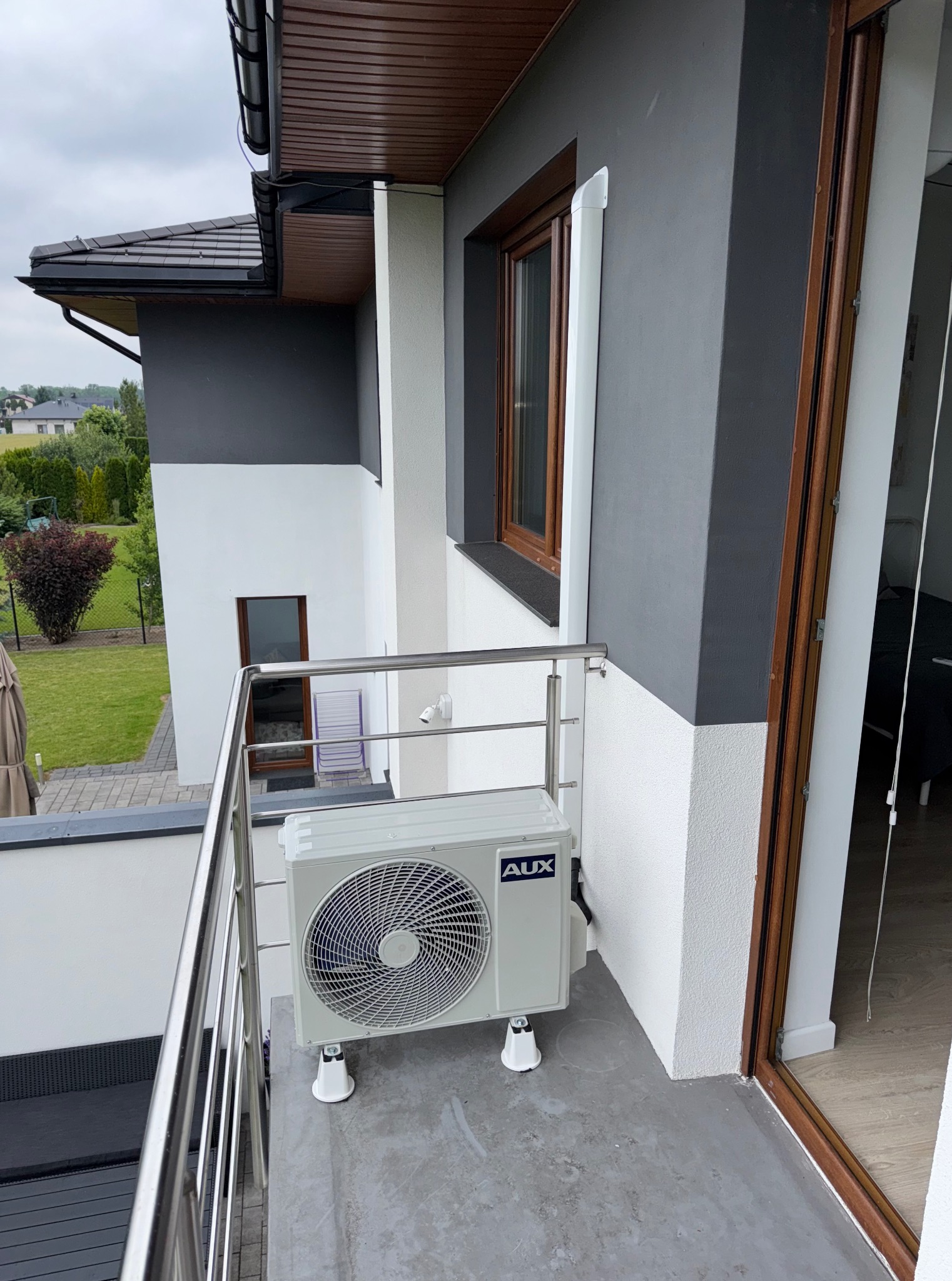 Biała jednostka zewnętrzna klimatyzacji AUX na balkonie nowoczesnego domu z szarą elewacją i drewnianymi oknami. Widok z góry na urządzenie i otoczenie.