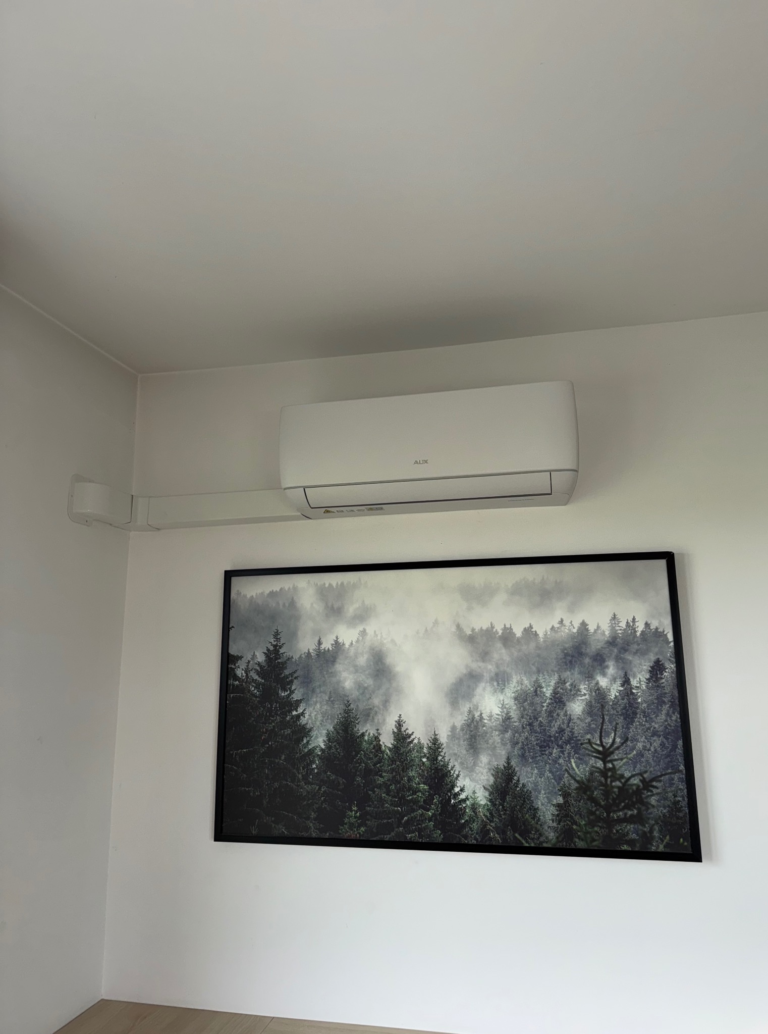Biała klimatyzacja AUX zamontowana na ścianie nad obrazem lasu we mgle w czarnej ramie. Widoczne białe rury instalacyjne. Minimalistyczny design wnętrza.