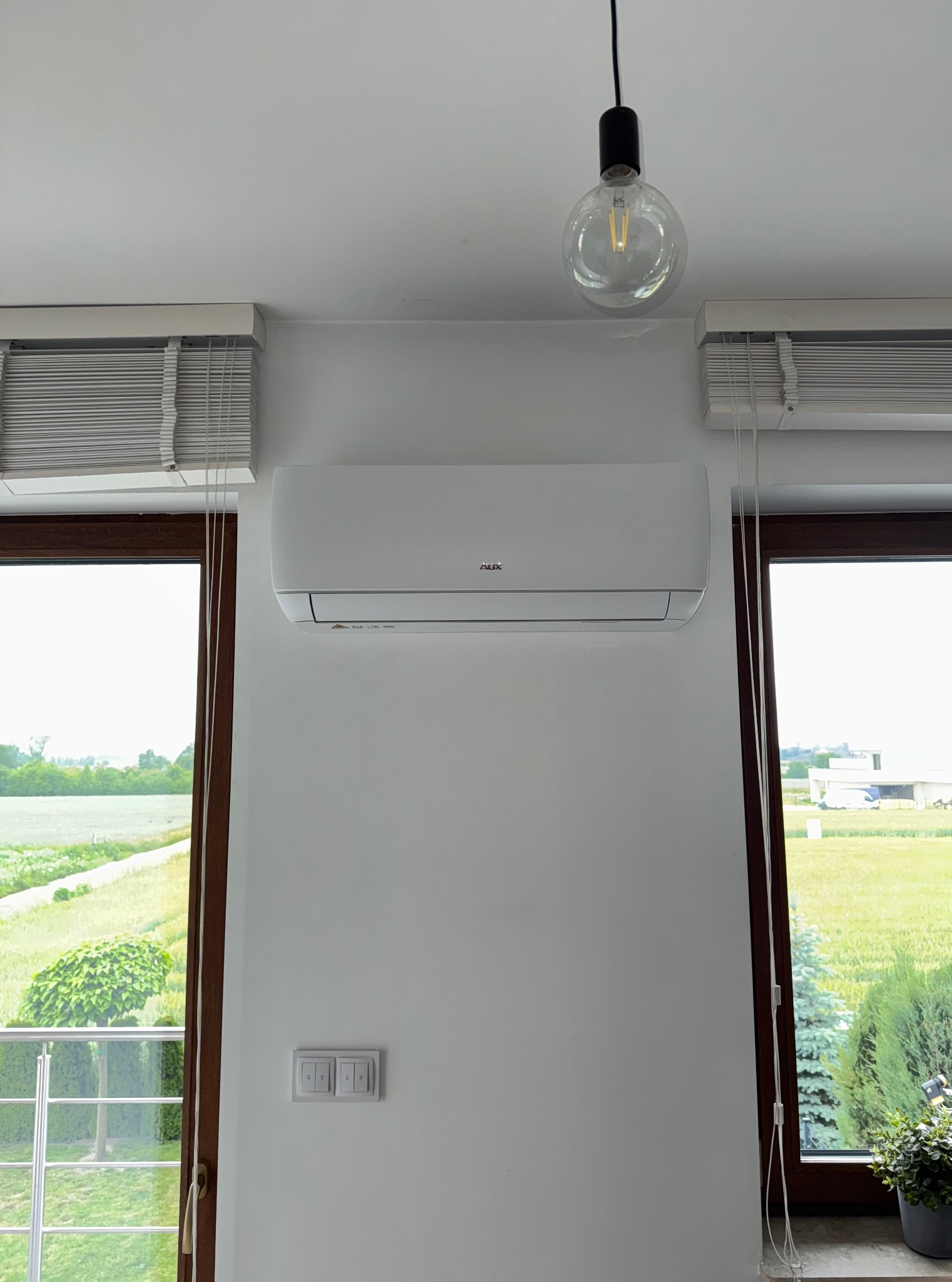 Biały klimatyzator AUX na ścianie w jasnym pokoju z widokiem na zieleń za oknem. Nad nim wisi żarówka dekoracyjna, a obok okien widoczne są białe żaluzje.