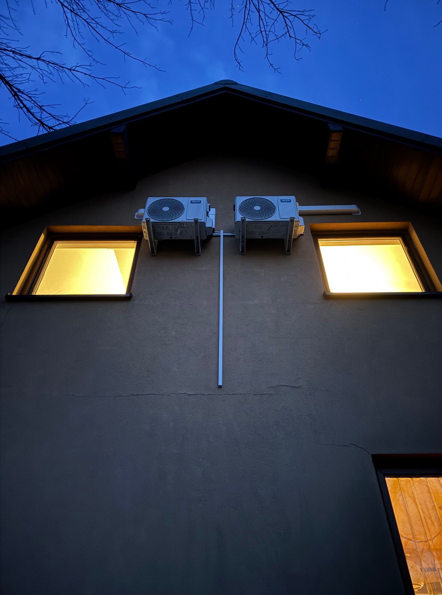 Dwa klimatyzatory AUX zamontowane symetrycznie nad oknami domu oświetlonymi ciepłym światłem, widoczne na tle wieczornego nieba i gałęzi drzew.