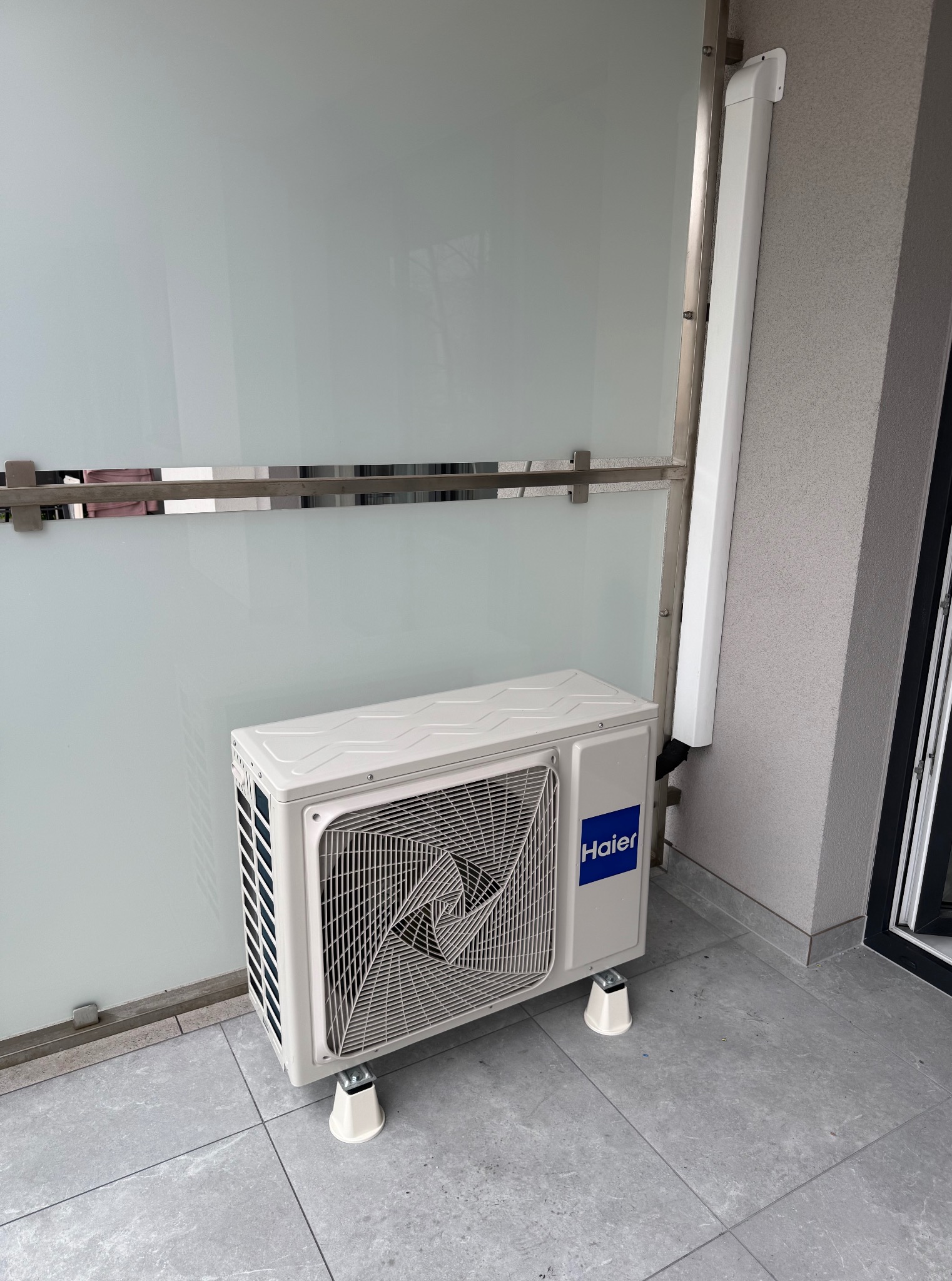 Biały klimatyzator Haier zamontowany na balkonie z szarą podłogą i szklaną balustradą. Widoczna rura odprowadzająca skropliny. Ujęcie z bliska.