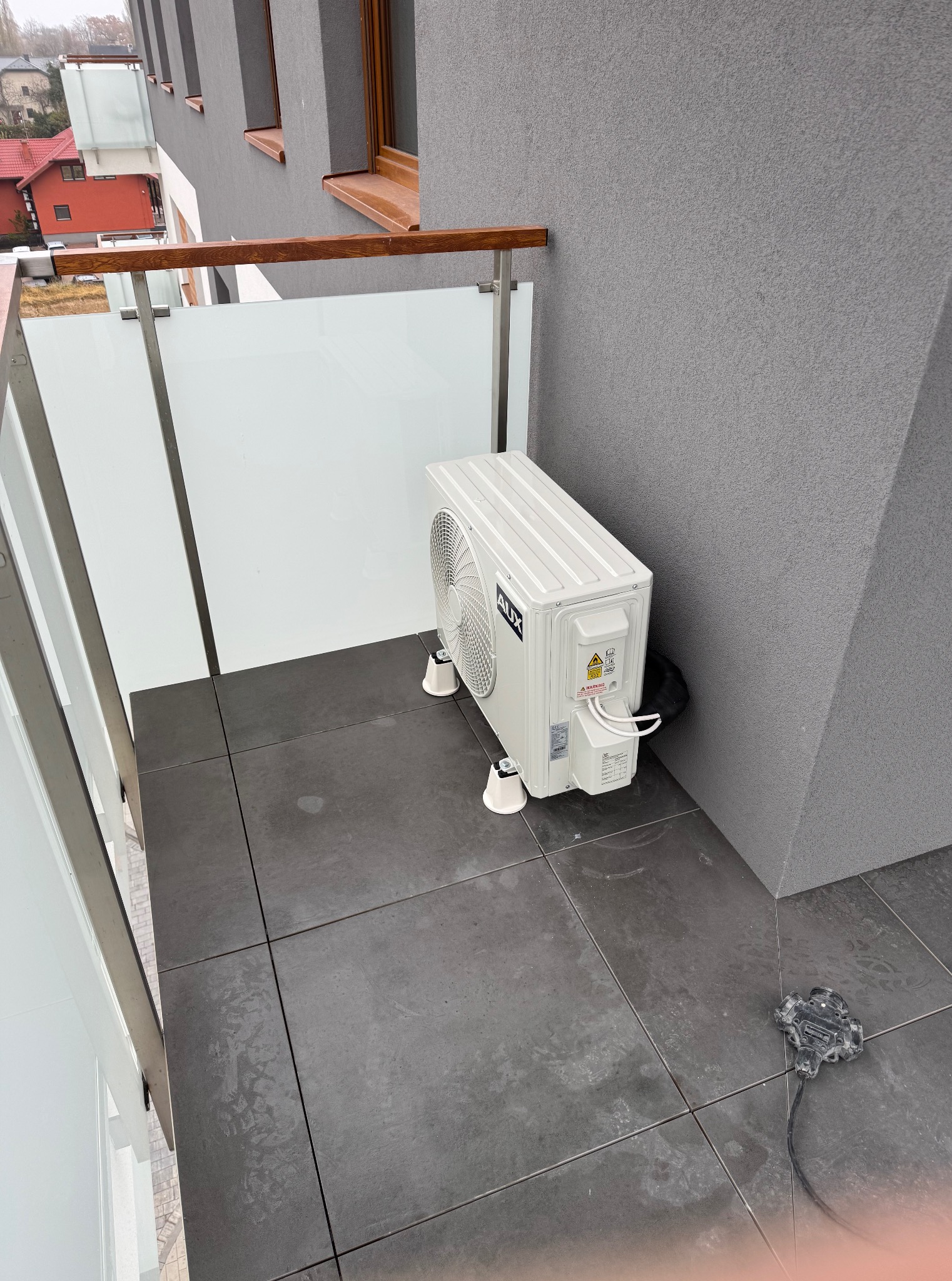 Biała jednostka zewnętrzna klimatyzacji AUX na balkonie z szarymi płytkami i szklaną balustradą. Widoczny kabel zasilający i fragment budynku w tle.
