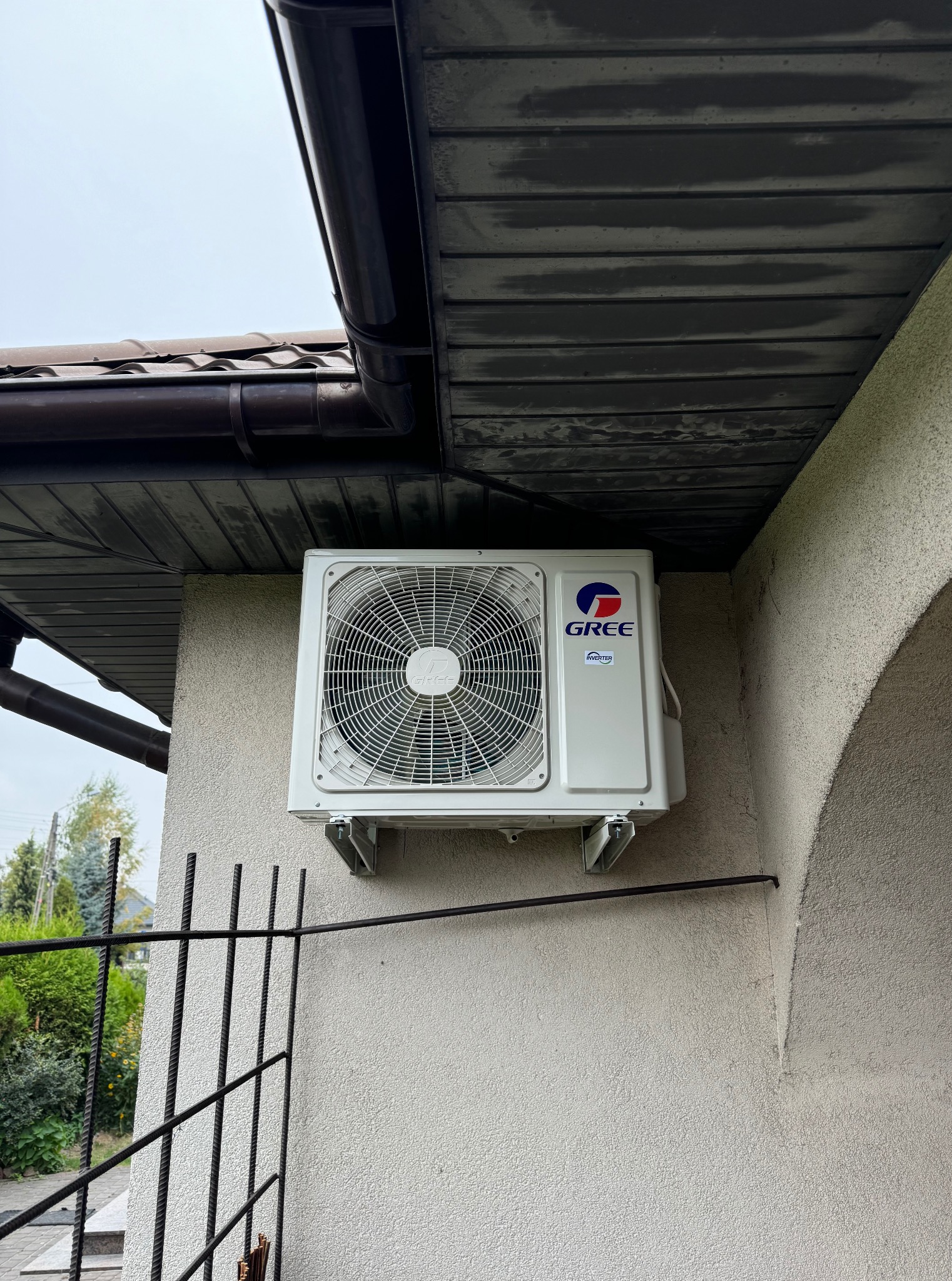 Biała jednostka zewnętrzna klimatyzacji Gree zamontowana na elewacji budynku, widoczna z dołu, z fragmentem rynny i ogrodzenia w kadrze.