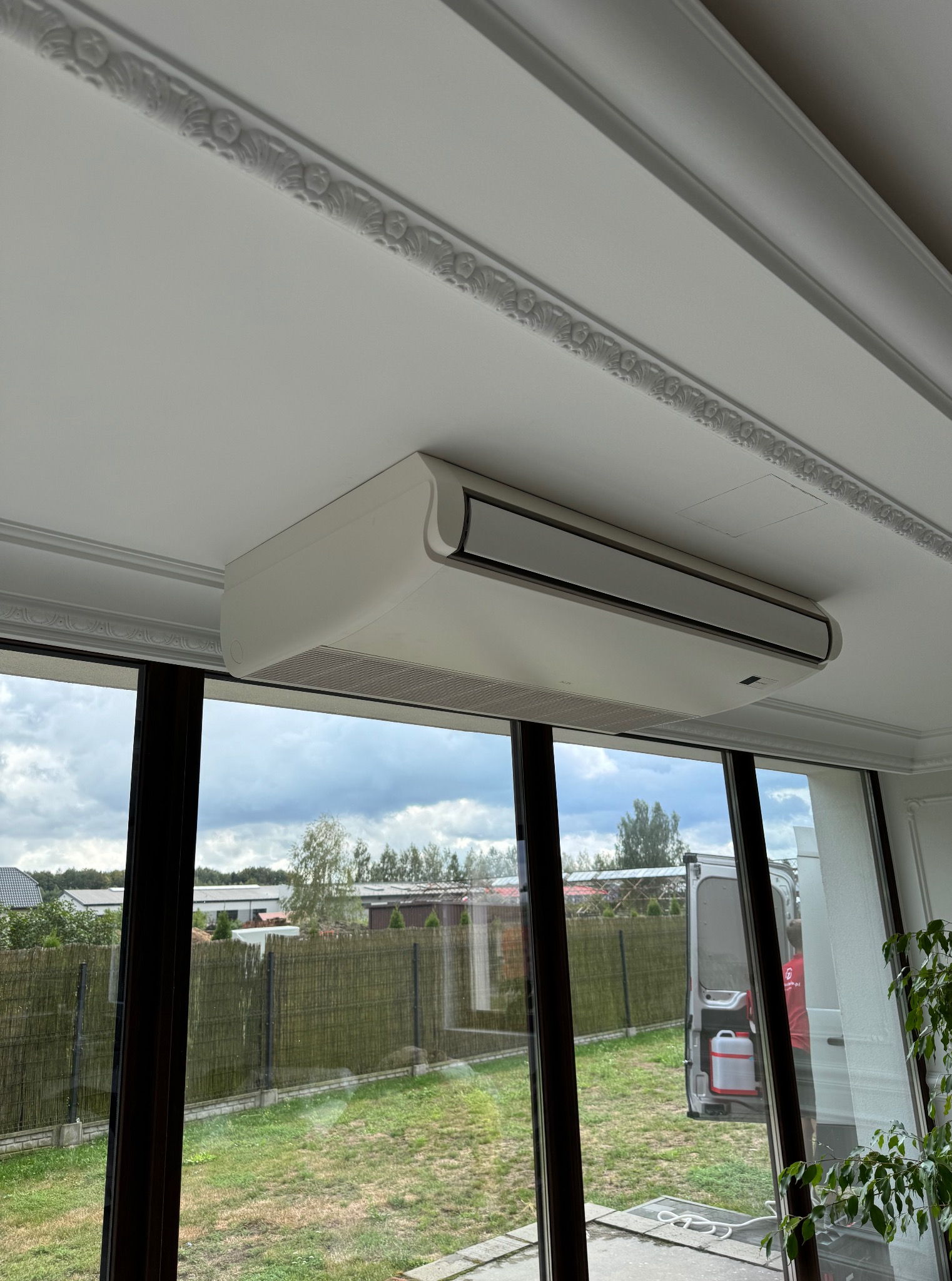 Biała klimatyzacja kasetonowa zamontowana pod sufitem z dekoracyjną sztukaterią, widok przez okno na zewnątrz. Częściowo widoczny samochód dostawczy.