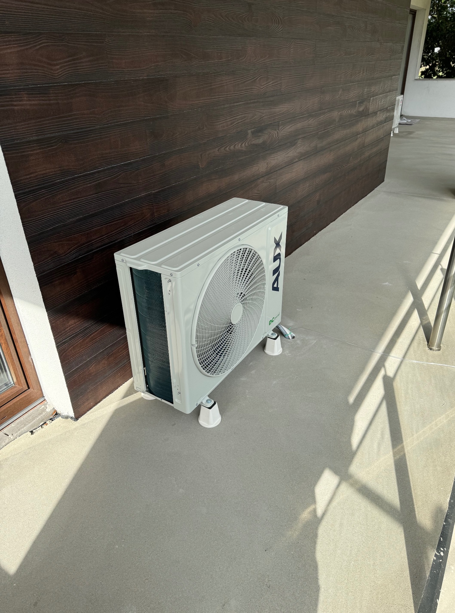 Biała jednostka zewnętrzna klimatyzacji AUX zamontowana na betonowym tarasie przy brązowej, drewnianej ścianie. Widoczne przewody i cień barierki.