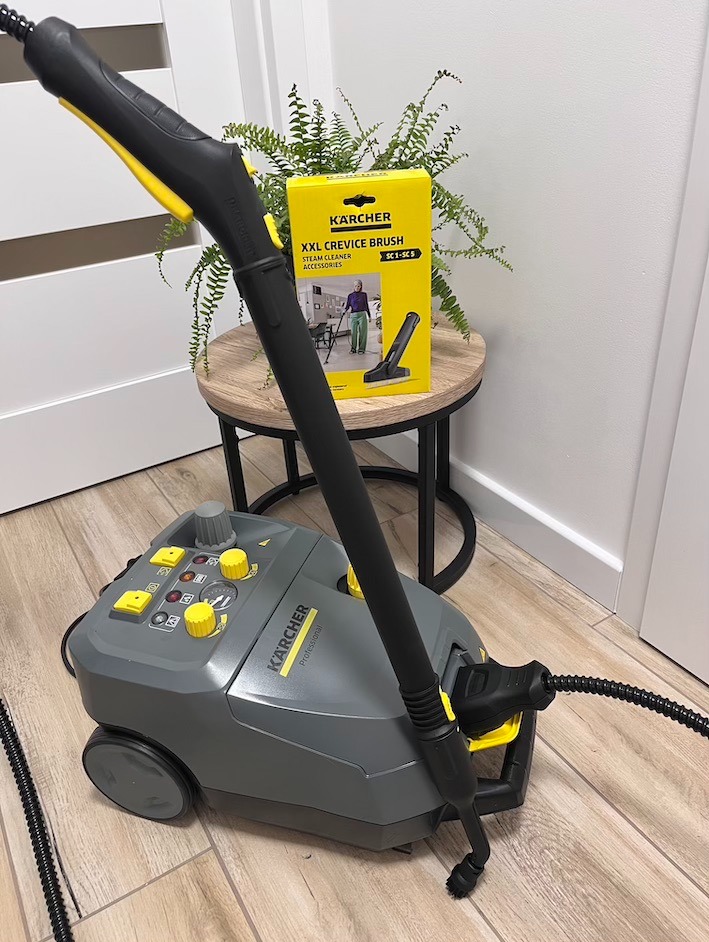 Profesjonalny parowy odkurzacz Kärcher z akcesoriami na drewnianej podłodze, obok stolika z rośliną i pudełkiem szczotki szczelinowej XXL.