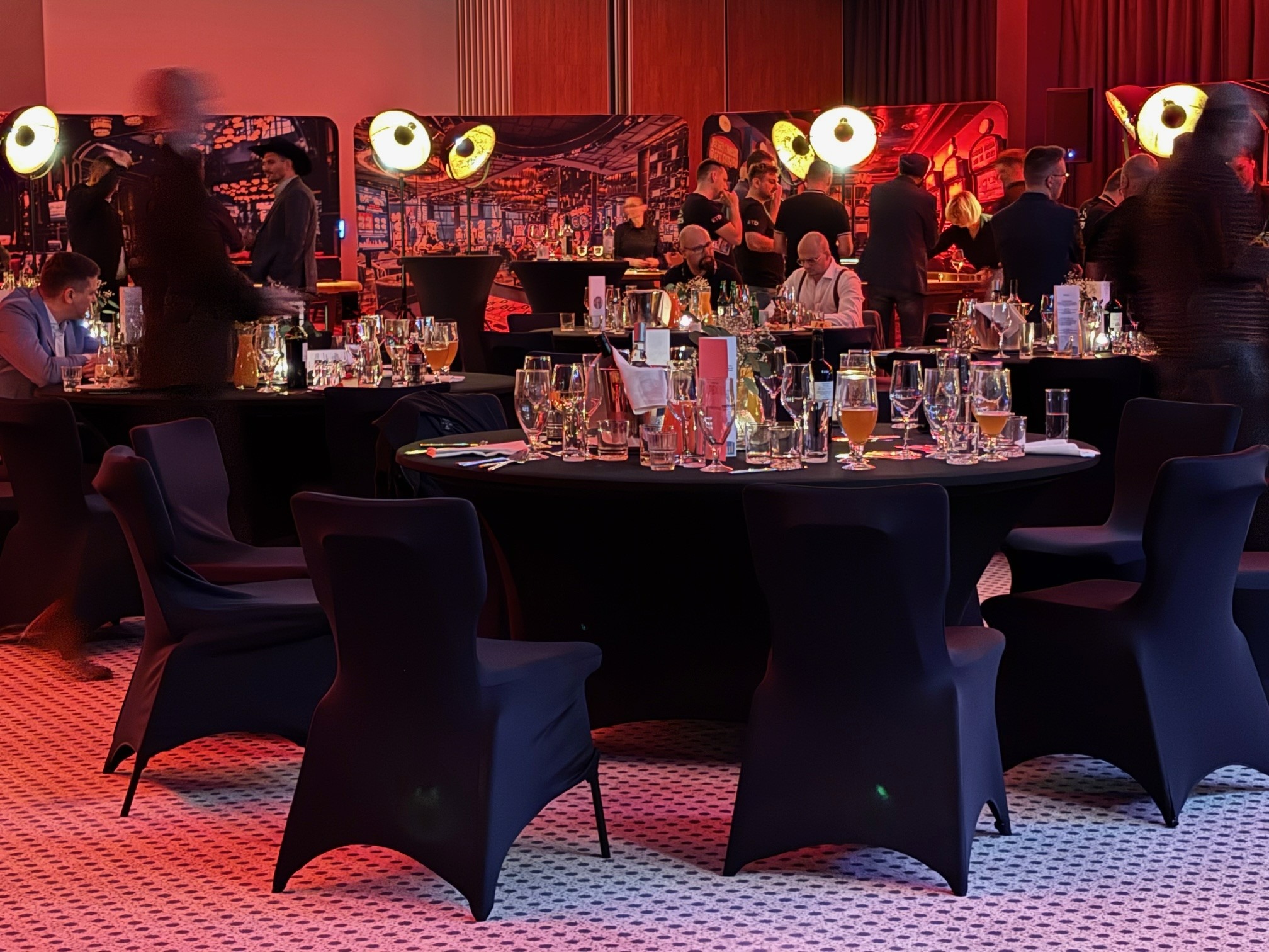 Ustawione stoły z czarnymi obrusami i krzesłami na sali bankietowej, gotowe na przyjęcie gości. Dekoracje świetlne i grafika w tle tworzą unikalny klimat.