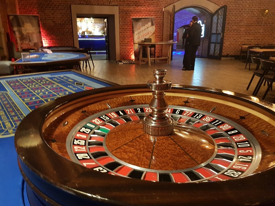 Kasyno eventowe: ruletka z drewnianym wykończeniem na pierwszym planie, w tle stoły do gier i osoba w garniturze w ceglanym wnętrzu.