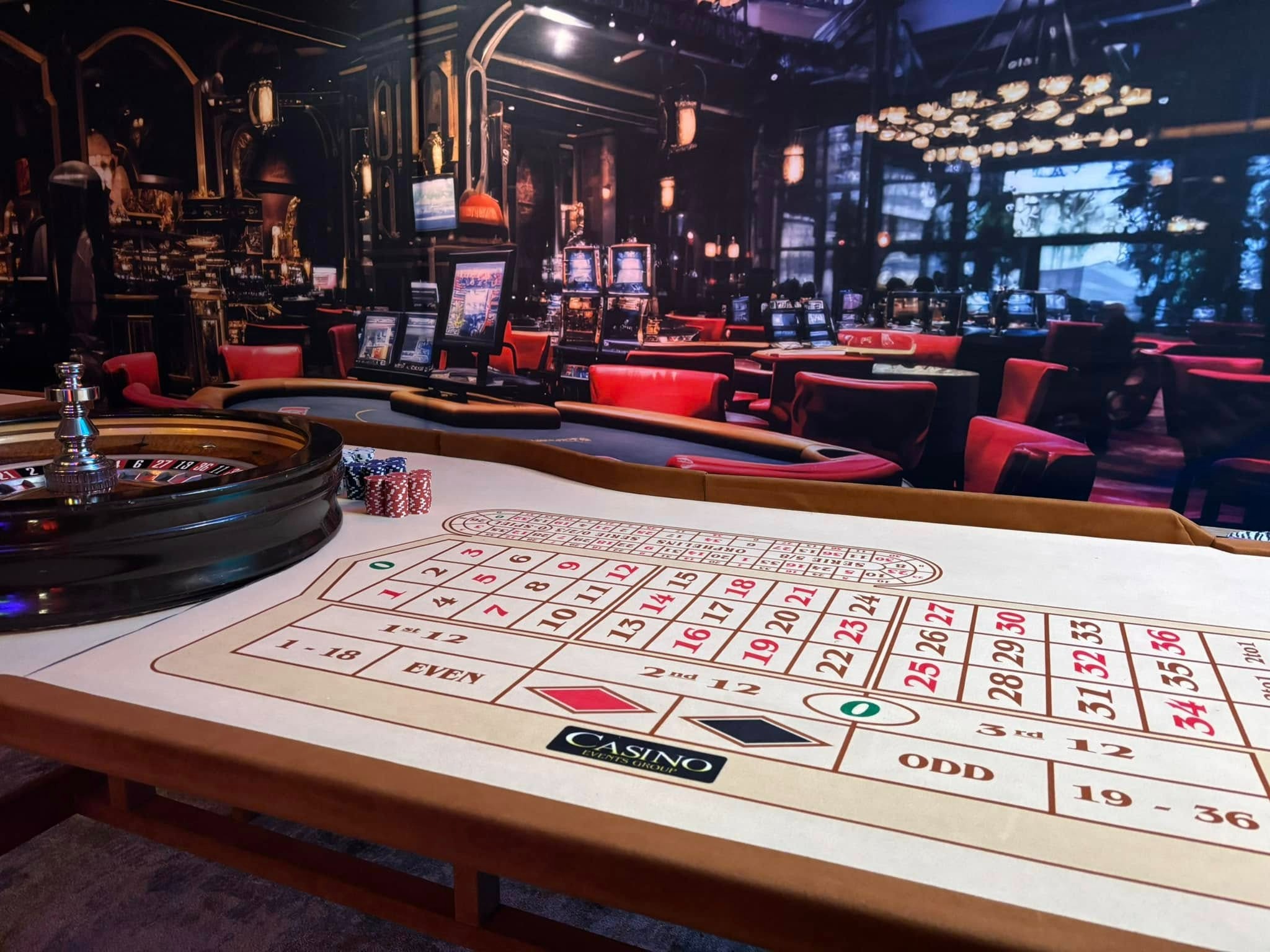 Stół do ruletki z żetonami i kołem, w tle kasyno z automatami i czerwonymi krzesłami. Kasyno Events Group.